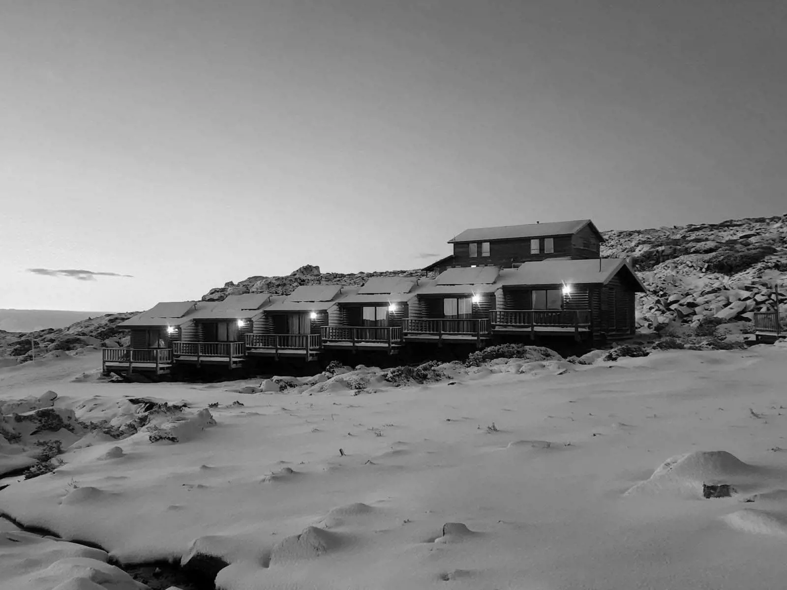 A row of cabins is illuminated at dusk, surrounded by a blanket of snow on a remote hillside.