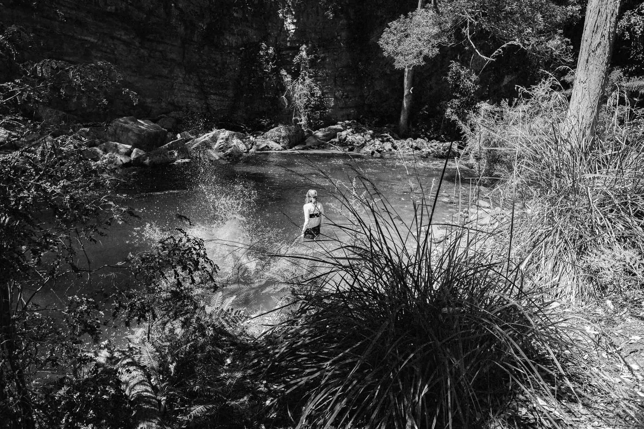 A person stands in shallow water at a rocky swimming spot, creating splashes around them.