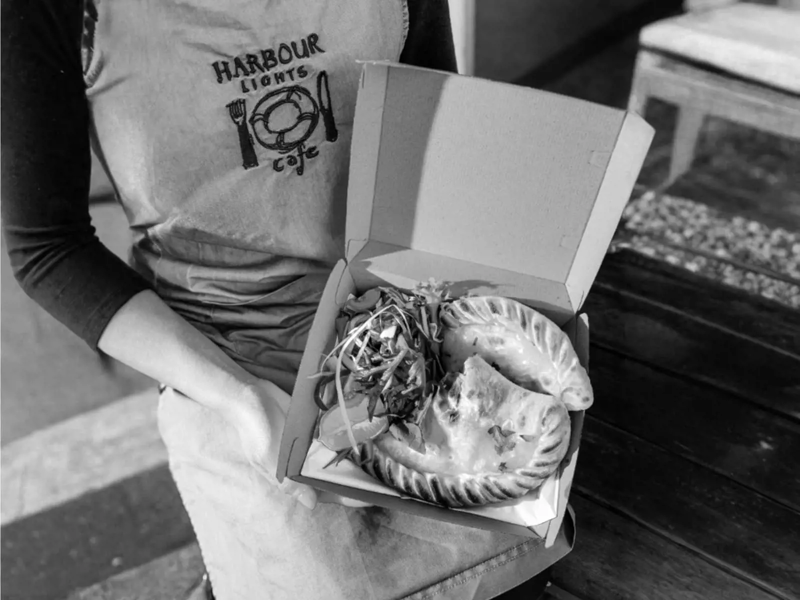 A person in an apron holds a takeout box containing two savory pastries and a side salad.