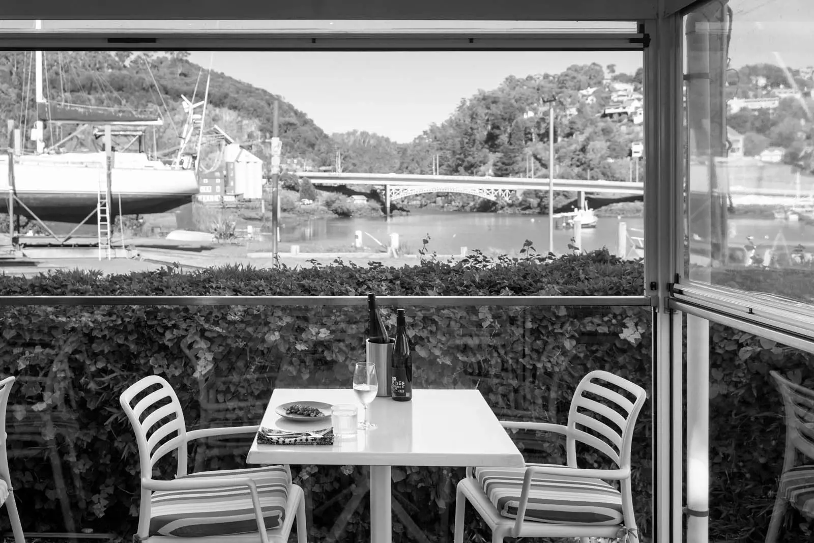 A table and two chairs in an outdoors undercover dining area, overlooking a river, a boatyard and a historic bridge.