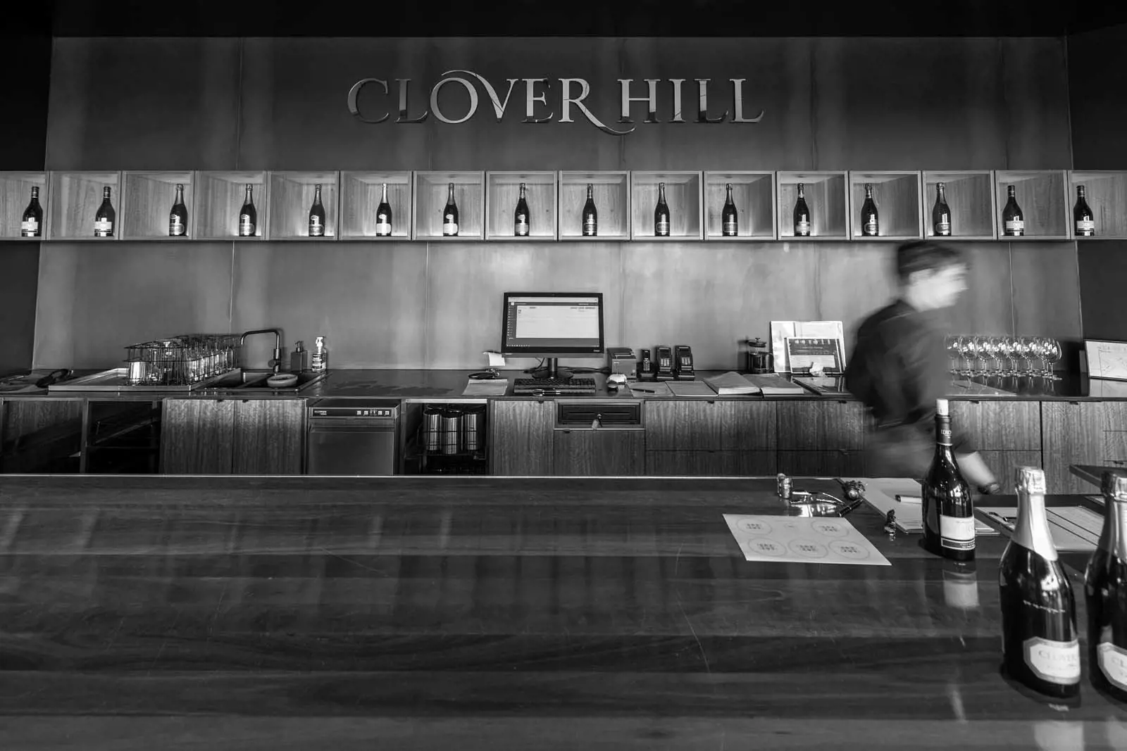 Looking over the smooth wooden bar of a winery, with bottles of sparkling lined artfully on the walls. The sign on the wall reads 'Clover Hill'.