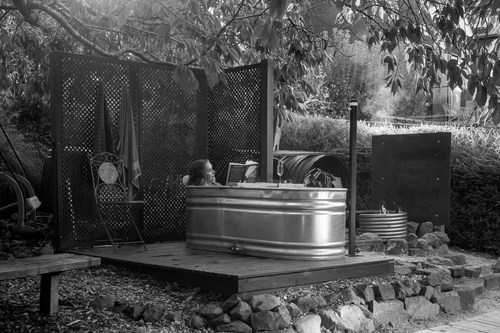A woman relaxes in an outdoor hot tub made from a galvanized steel container, reading a book with a glass of sparkling wine nearby. Surrounding her, lush greenery and decorative privacy panels create a tranquil and luxurious ambiance.