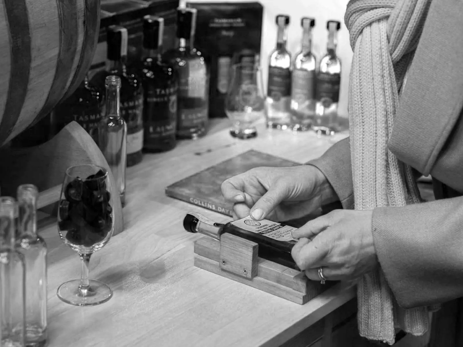 A person is carefully pasting a label onto a small bottle lying on a table. Other whiskies, bottles and glasses surround the worktop.