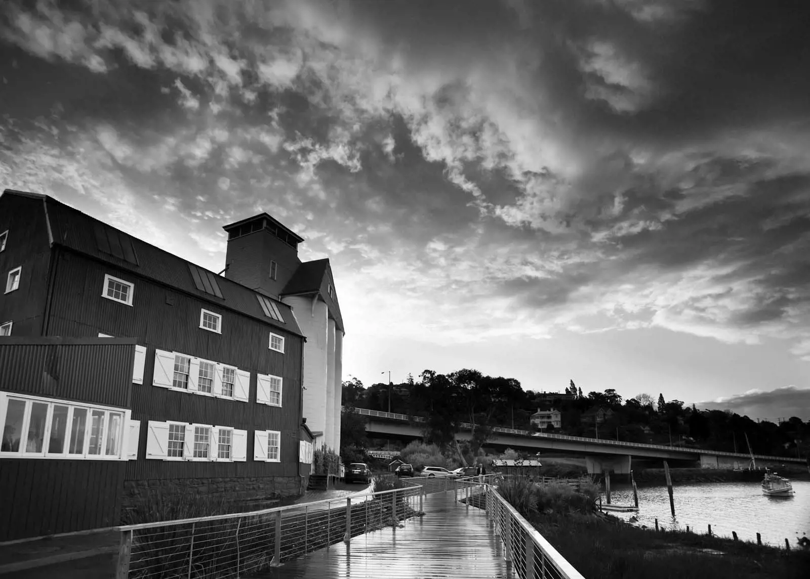 A three-storey building overlooks a picturesque river, with a boardwalk snaking in between. Heavy, atmospheric clouds hang in the sky above.