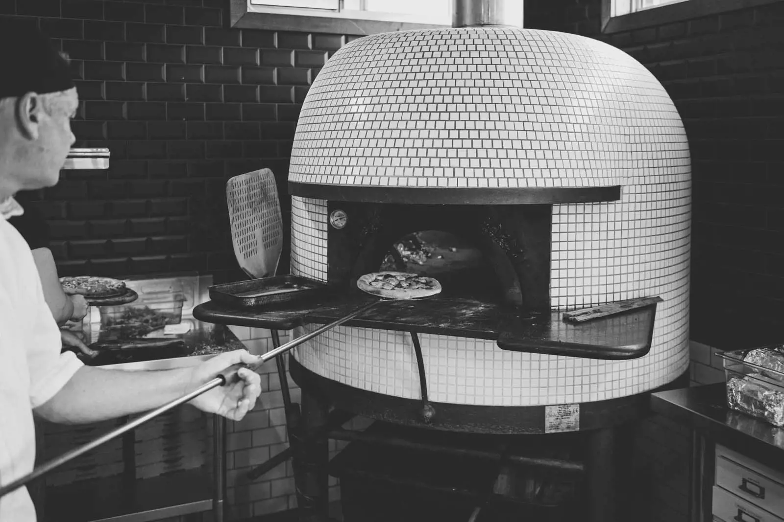 A chef uses a long-handled paddle to pull a cooked pizza from a domed, white-tiled woodfired pizza oven.