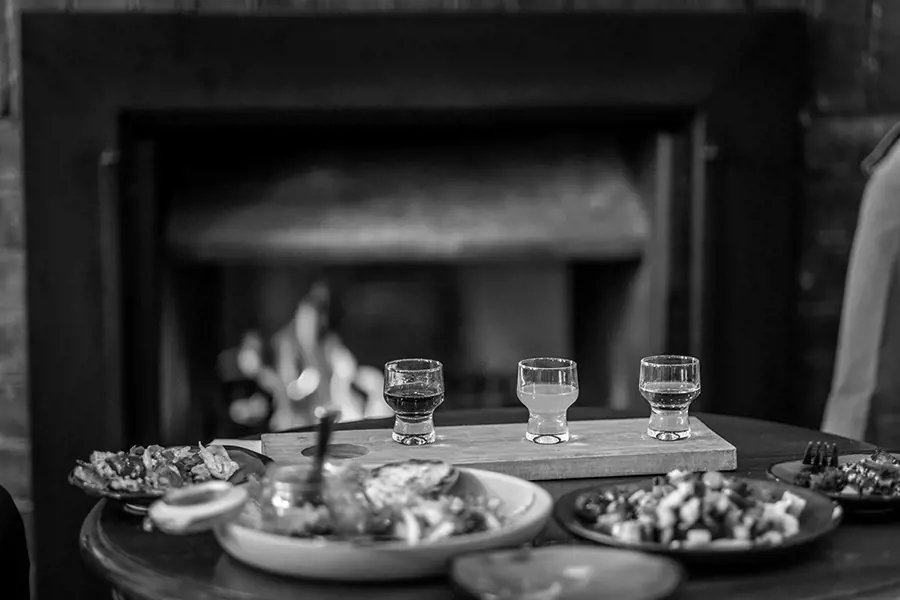 A rustic table set with hearty meals and a tasting paddle of drinks sits near a crackling fireplace, creating a warm and welcoming winter scene at the Empire Hotel.