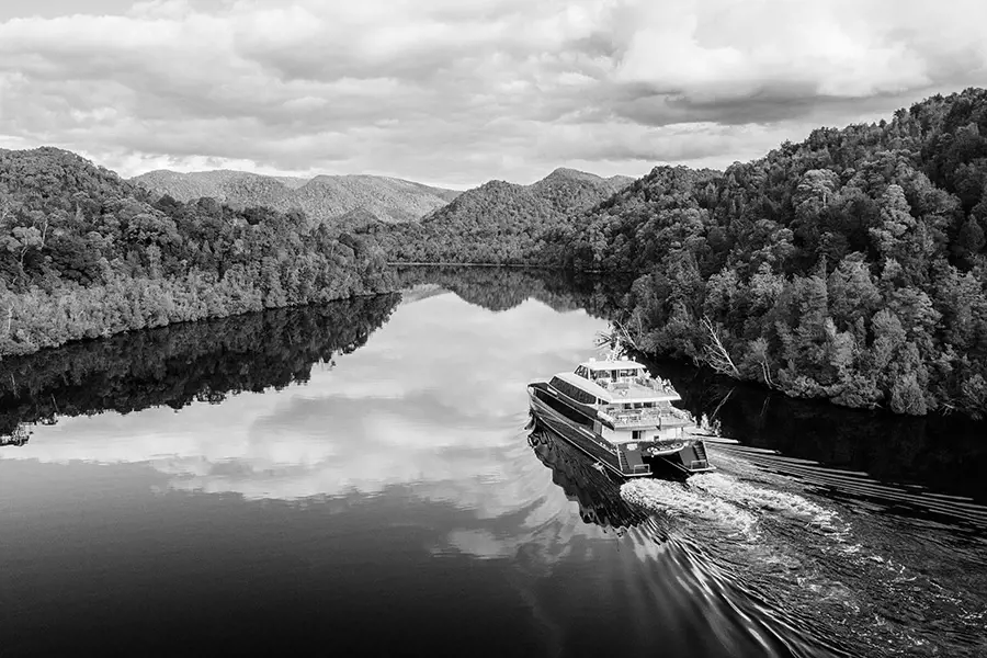 A cruise boat glides through the still, misty waters of Tasmania’s west coast wilderness under a grey winter sky.