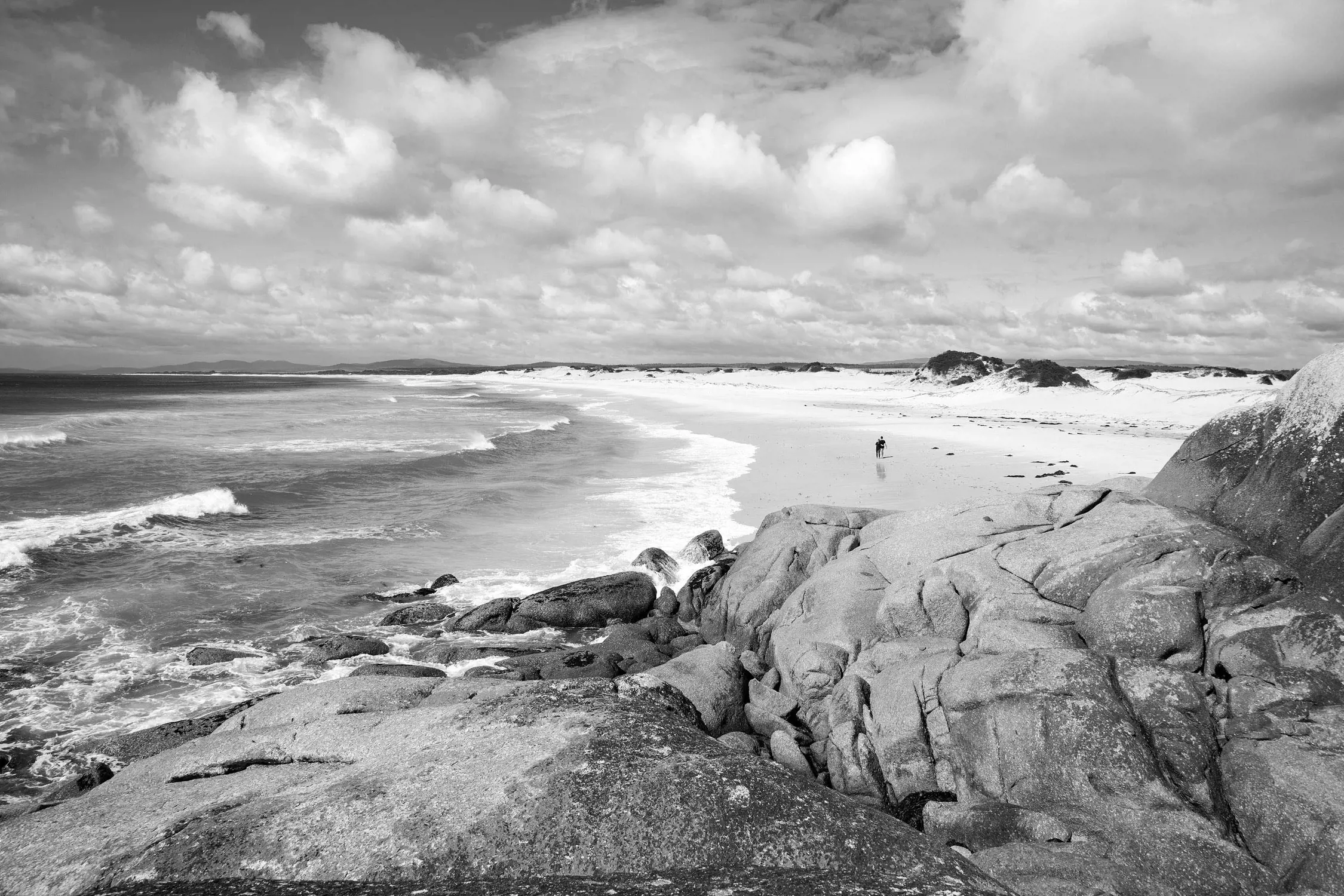 Panoramic view of a beach with waves, sandy shore, rocks in the foreground, and two people walking in the distance.