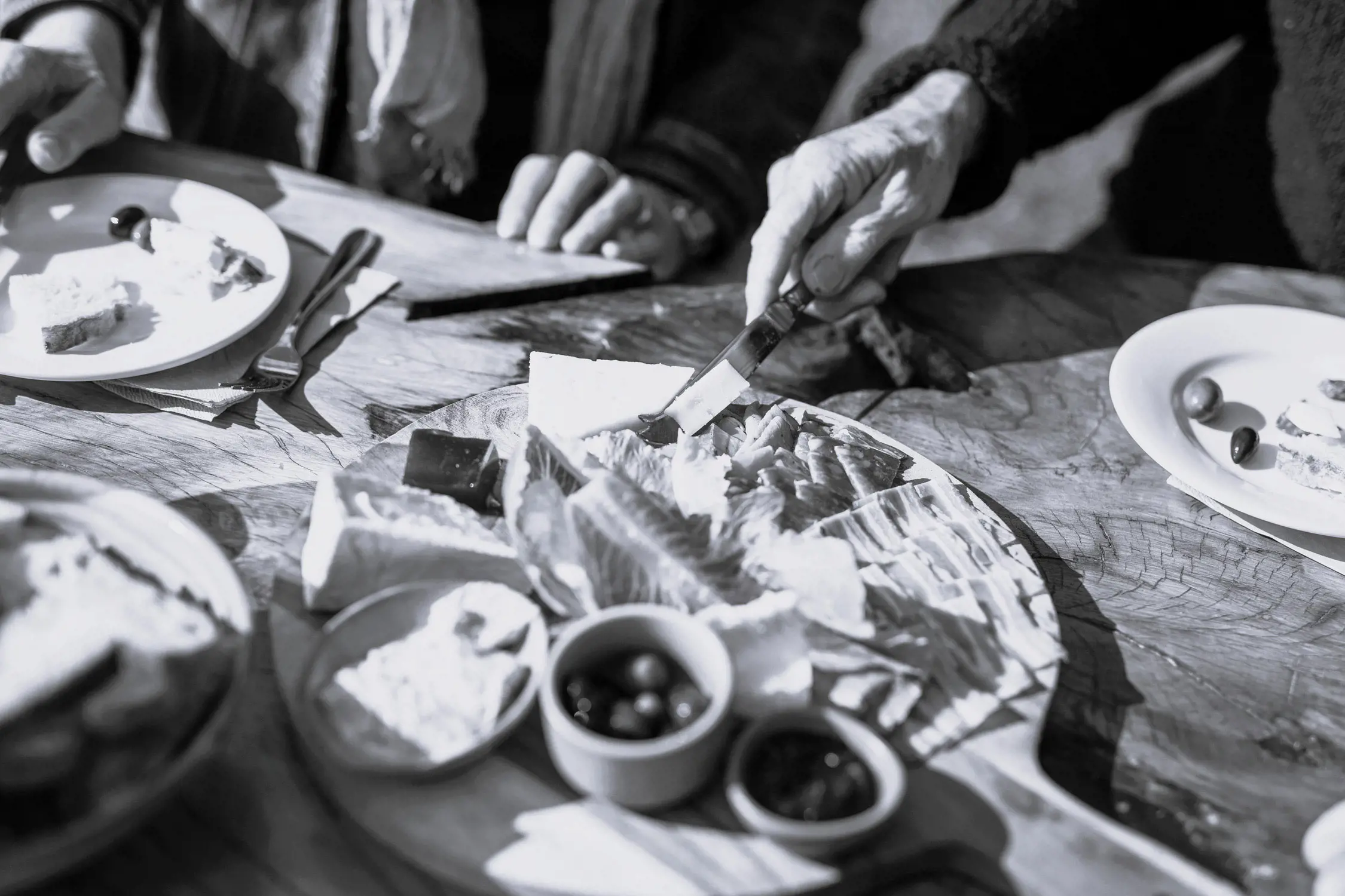 People gathered around a wooden table with a cheese board containing various cheeses, olives, and spreads.