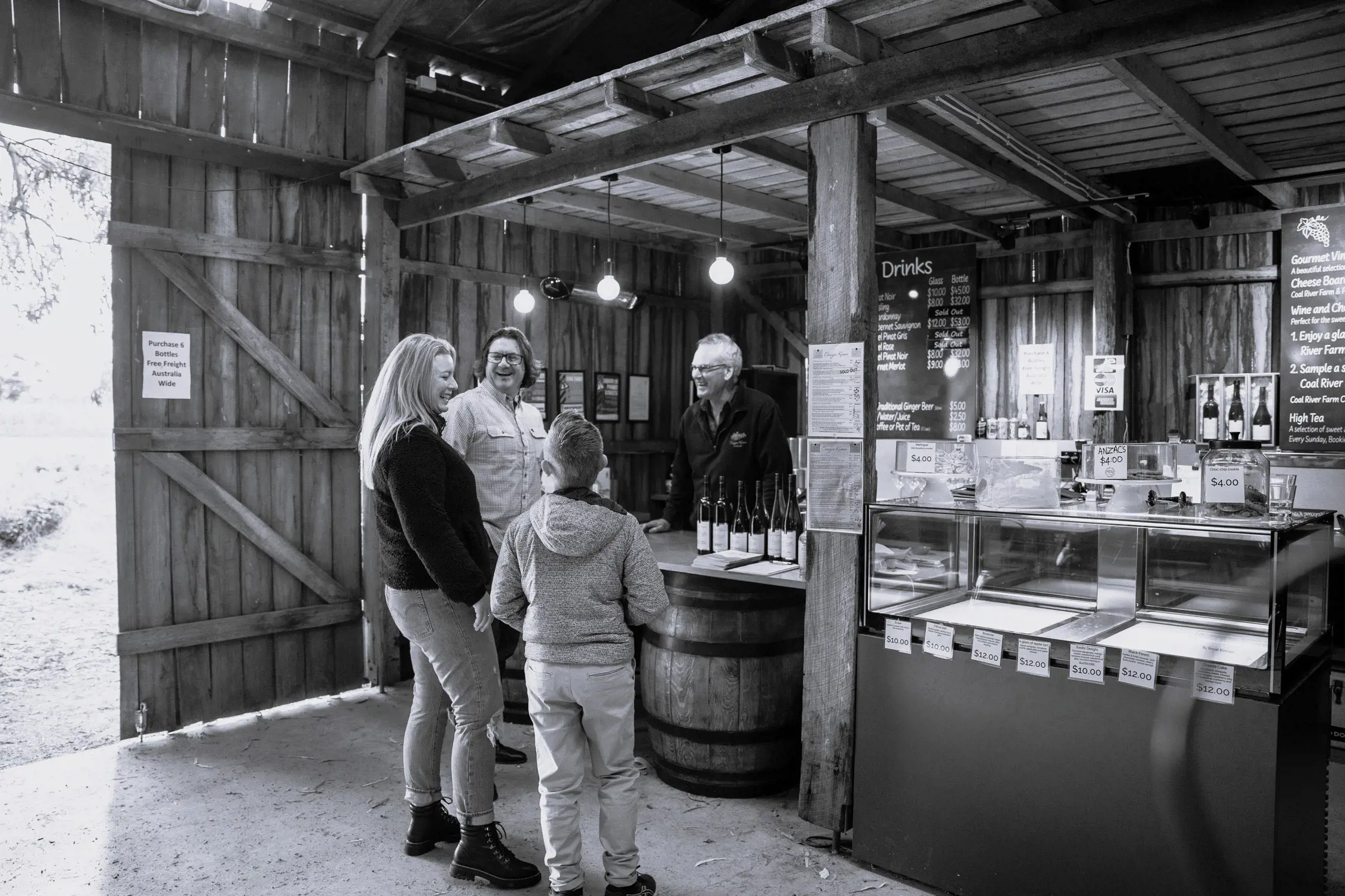 Inside a rustic, wooden wine shop, two adults and a child interact with a shopkeeper behind a counter displaying wine bottles and various items.