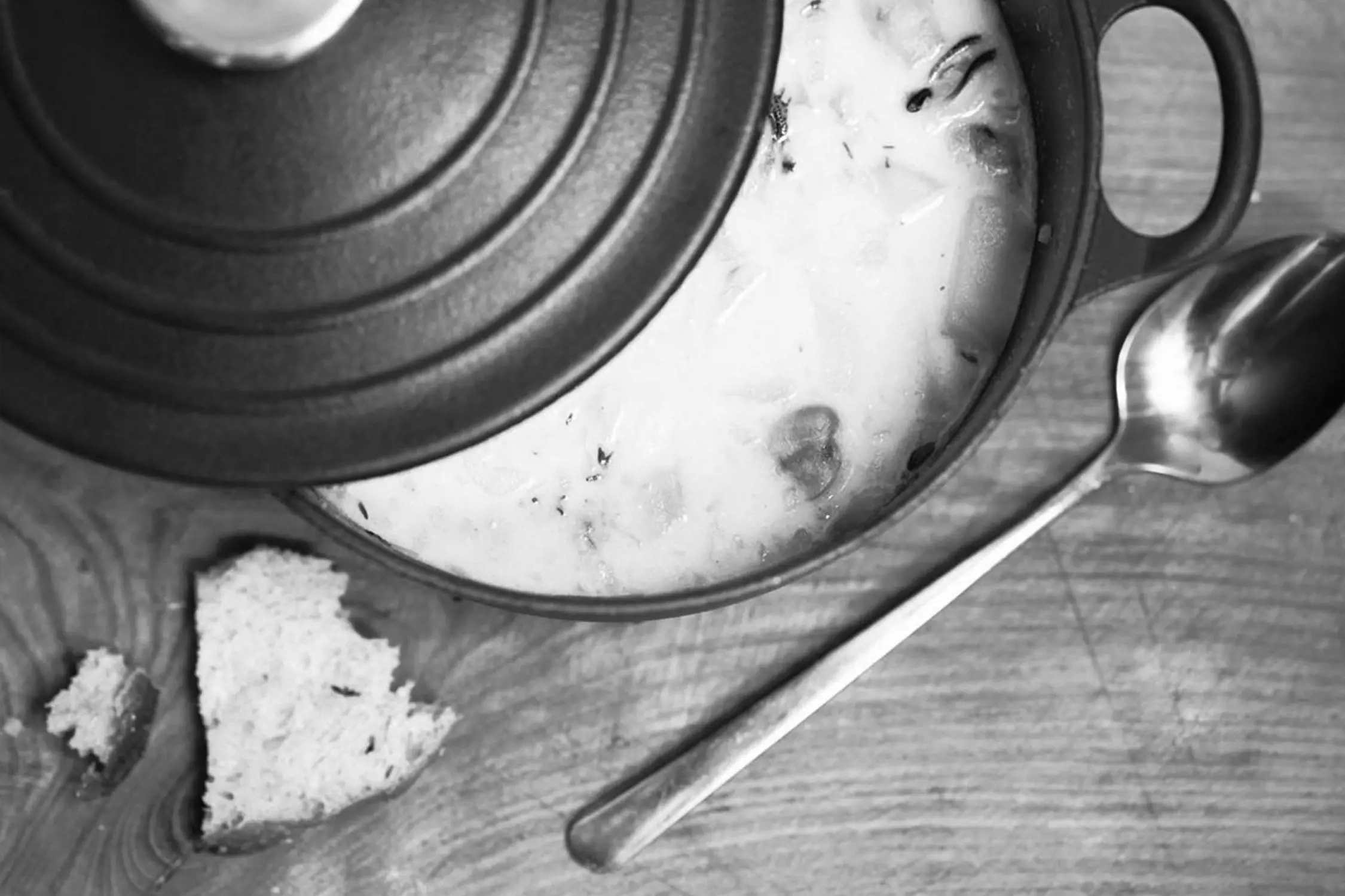 Close-up black and white image of a partially covered pot on a wooden surface containing a creamy chowder, with pieces of bread next to it and a spoon resting nearby.