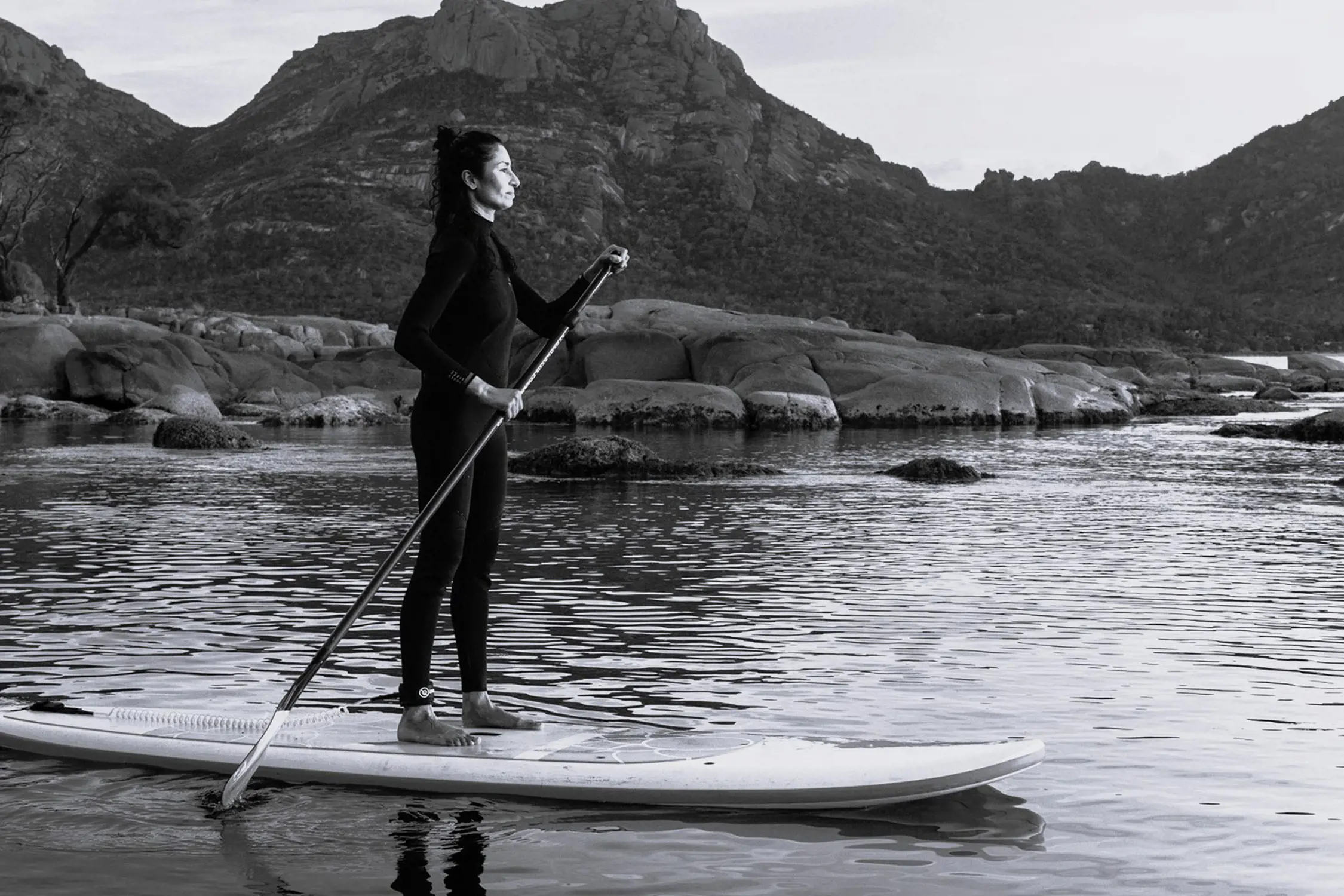 Black and white picture of a person standing on a paddleboard in calm waters near rocky terrain, holding a paddle and wearing a wetsuit, with mountains and trees in the background.