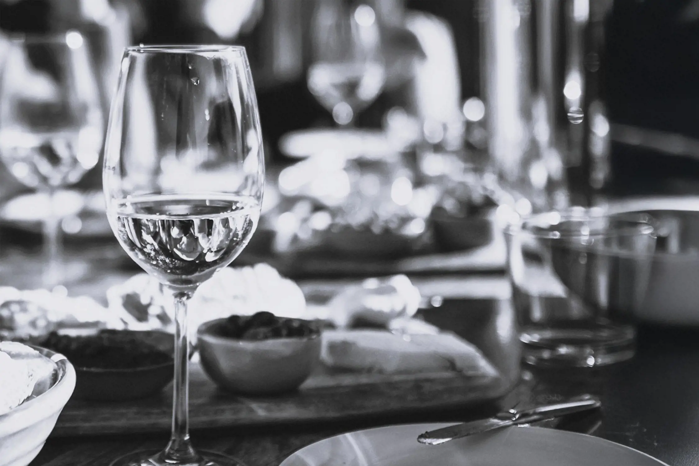 Black and white photo of a wine glass on a table, with blurred background showing more tableware and food items.