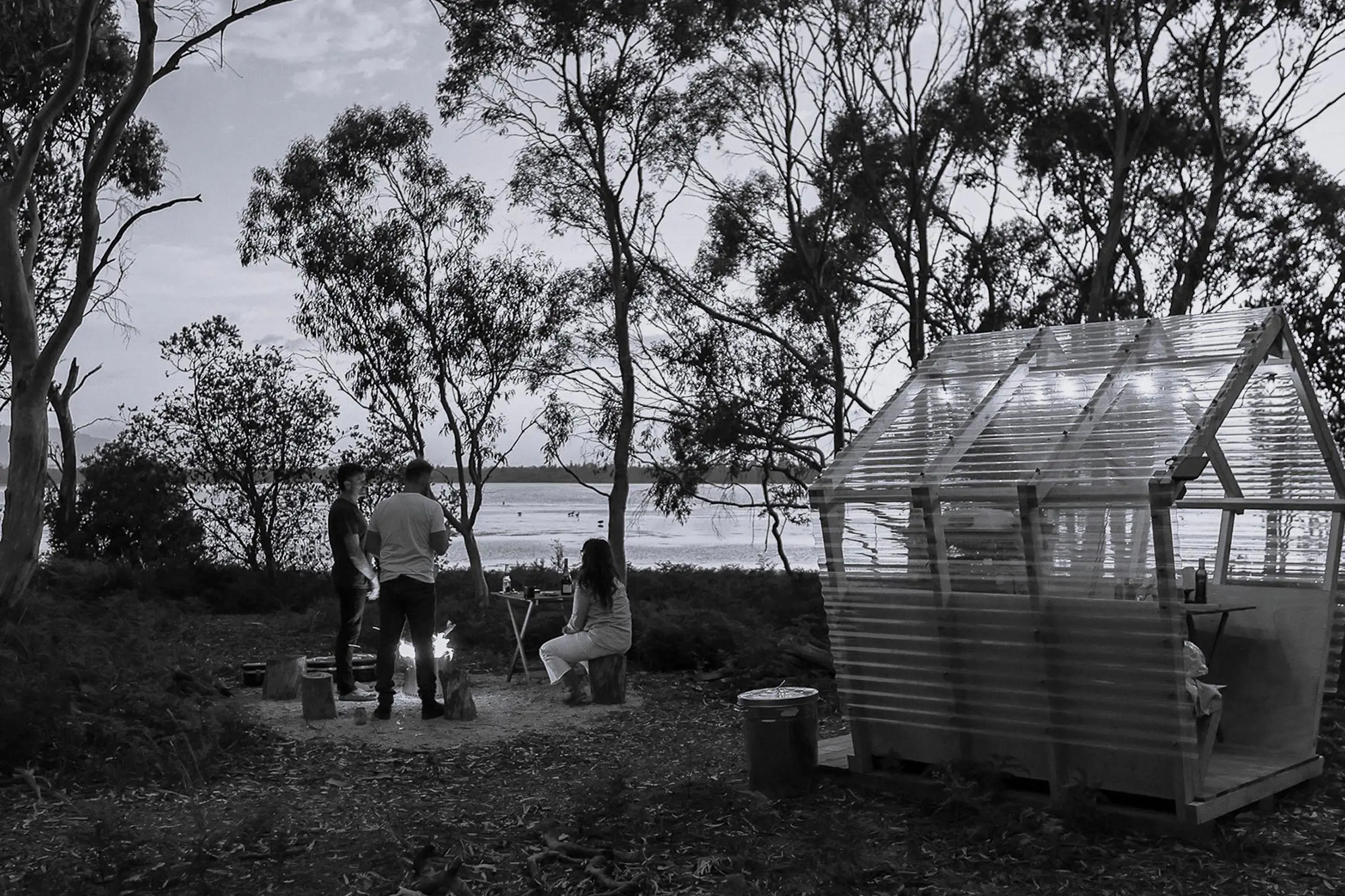 Black and white image of three people gathered near a fire pit in a wooded area next to a transparent small shelter, overlooking a body of water in the distance.
