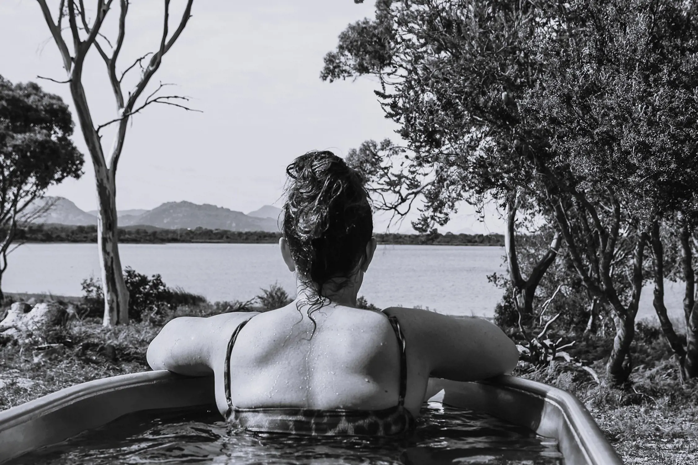 Black and white photo of a person with wet hair relaxing in an outdoor bathtub, facing away from the camera towards a lake and surrounded by trees.