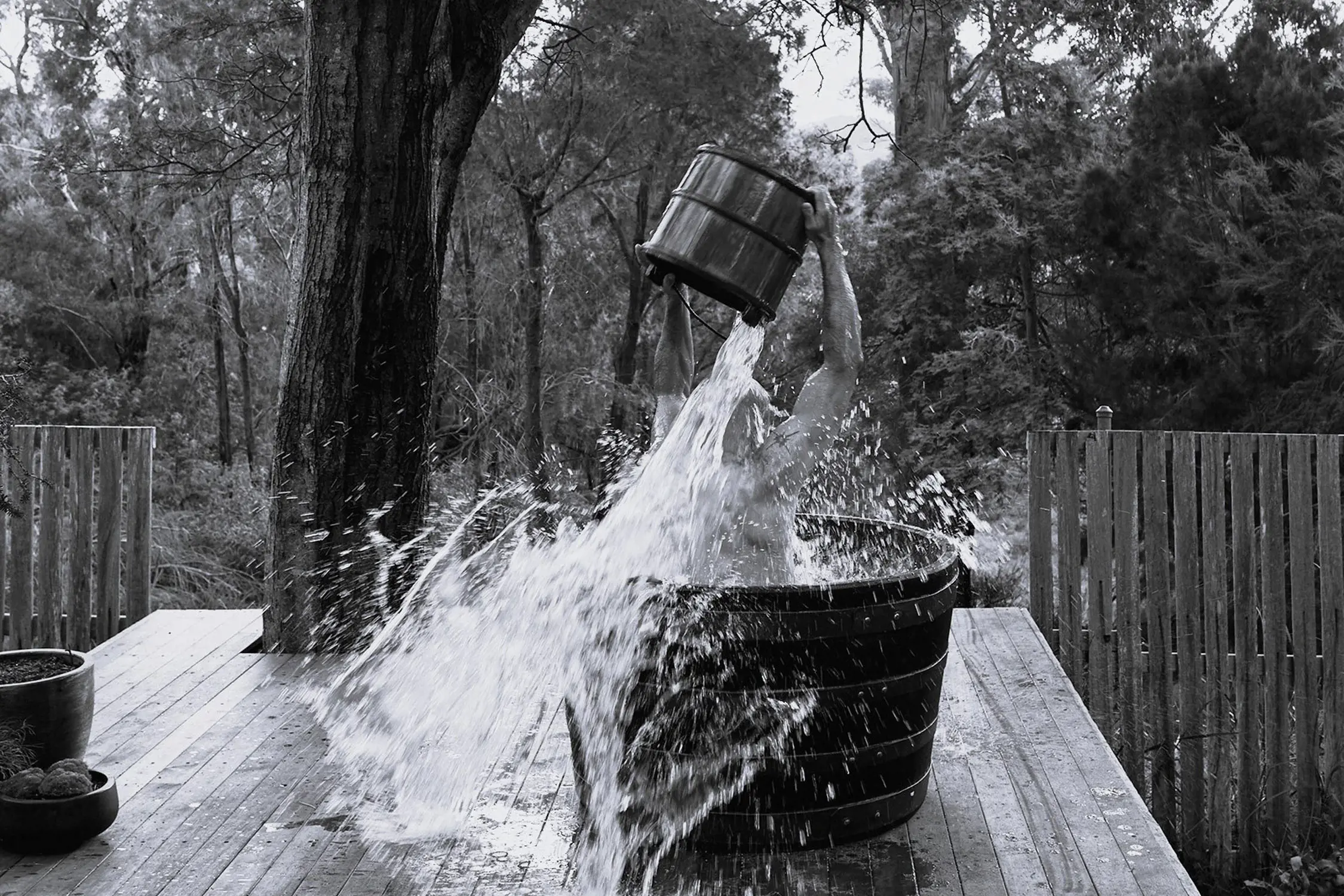 Black and white image of a person sitting in a large wooden tub on a wooden deck surrounded by trees, dumping a bucket of water over themselves with water splashing dramatically.