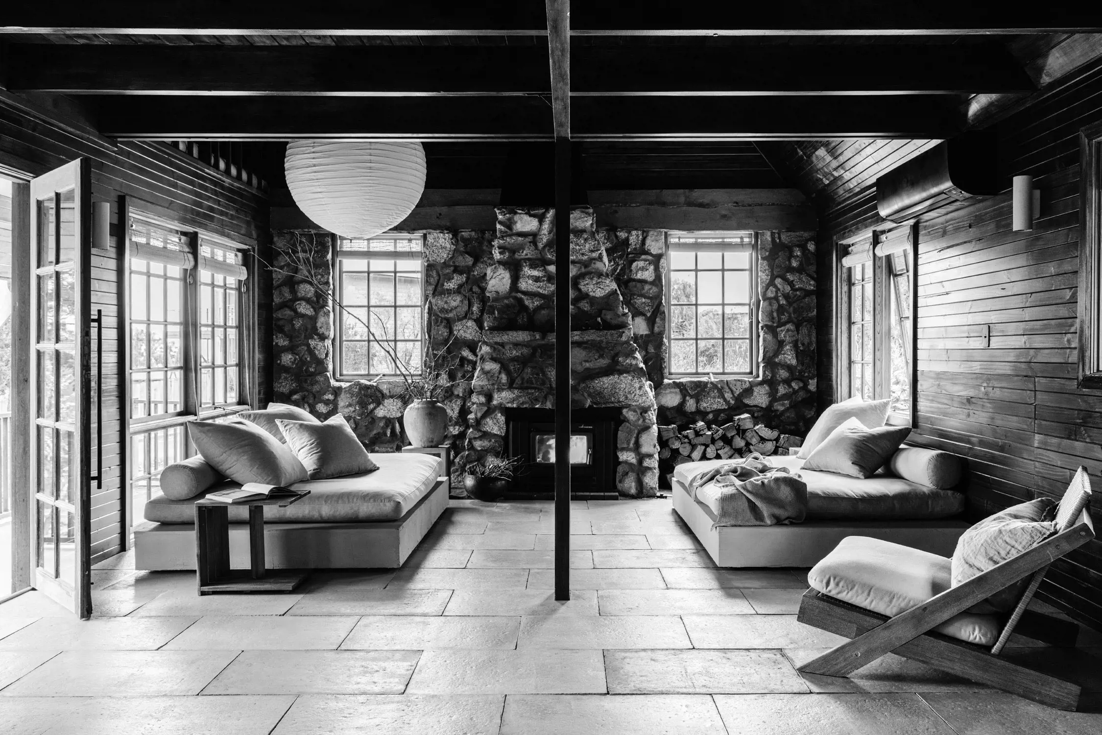 Black and white image of a cozy living room with two cushioned sofas facing each other, a stone fireplace centered between two windows, wood paneling on the walls, a lantern-style light hanging from the ceiling, and a side table next to one sofa.