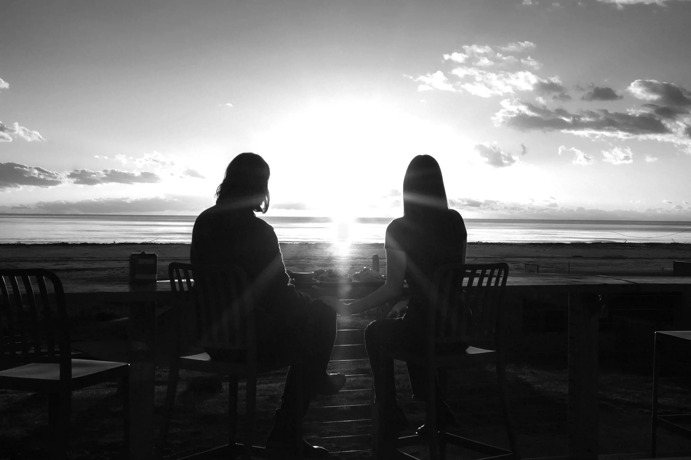 Black and white image of two people sitting on stools at a high table facing a beach and ocean at sunset or sunrise, silhouetted by the bright sun low on the horizon.