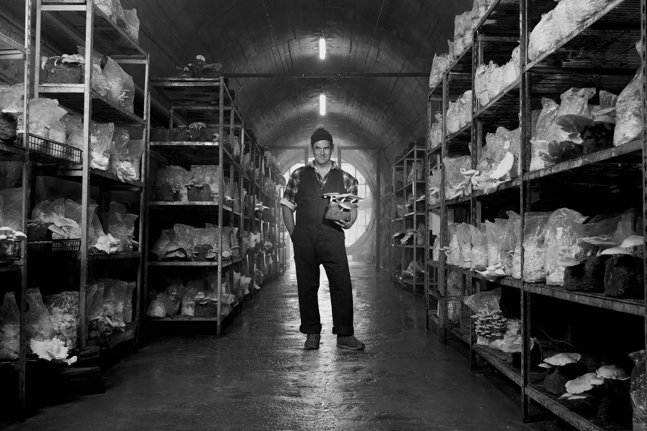 Black and white photo of a person wearing overalls and a hat standing in the middle of a mushroom growing facility, holding a cluster of mushrooms. Shelves filled with mushroom bags line both sides of the space.