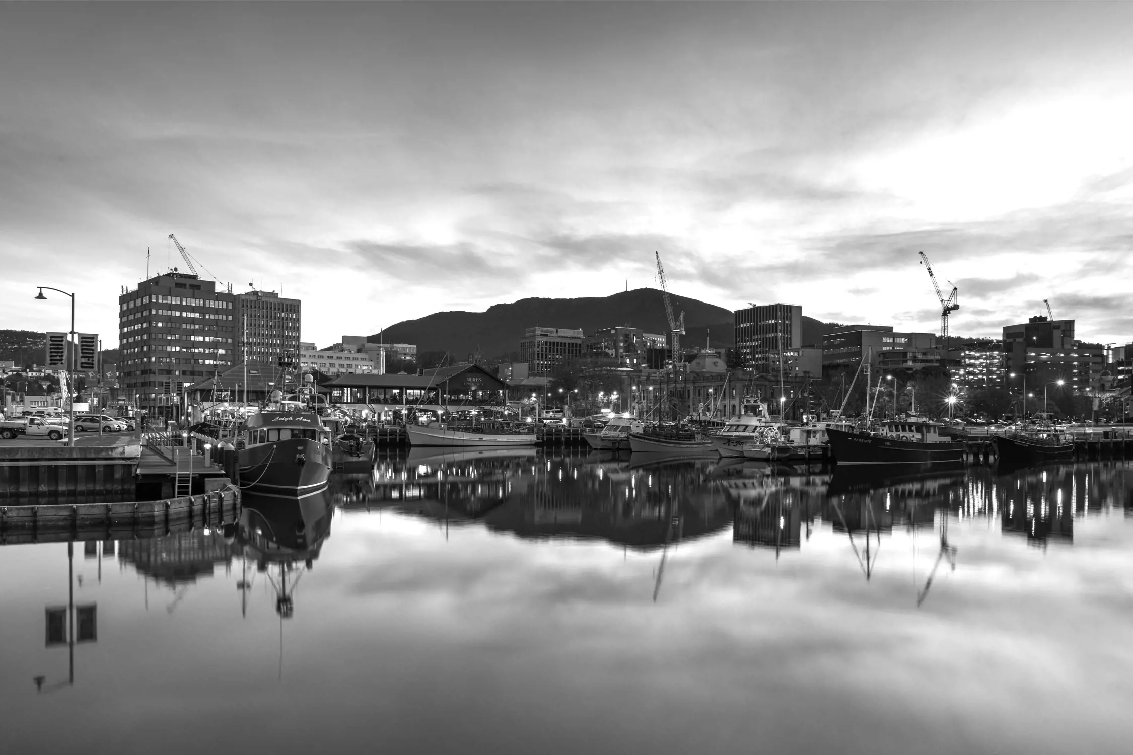 Black and white photo of a harbor with several boats docked, city buildings in the background, and a mountain behind the city. The water is calm, creating clear reflections of the boats and buildings.