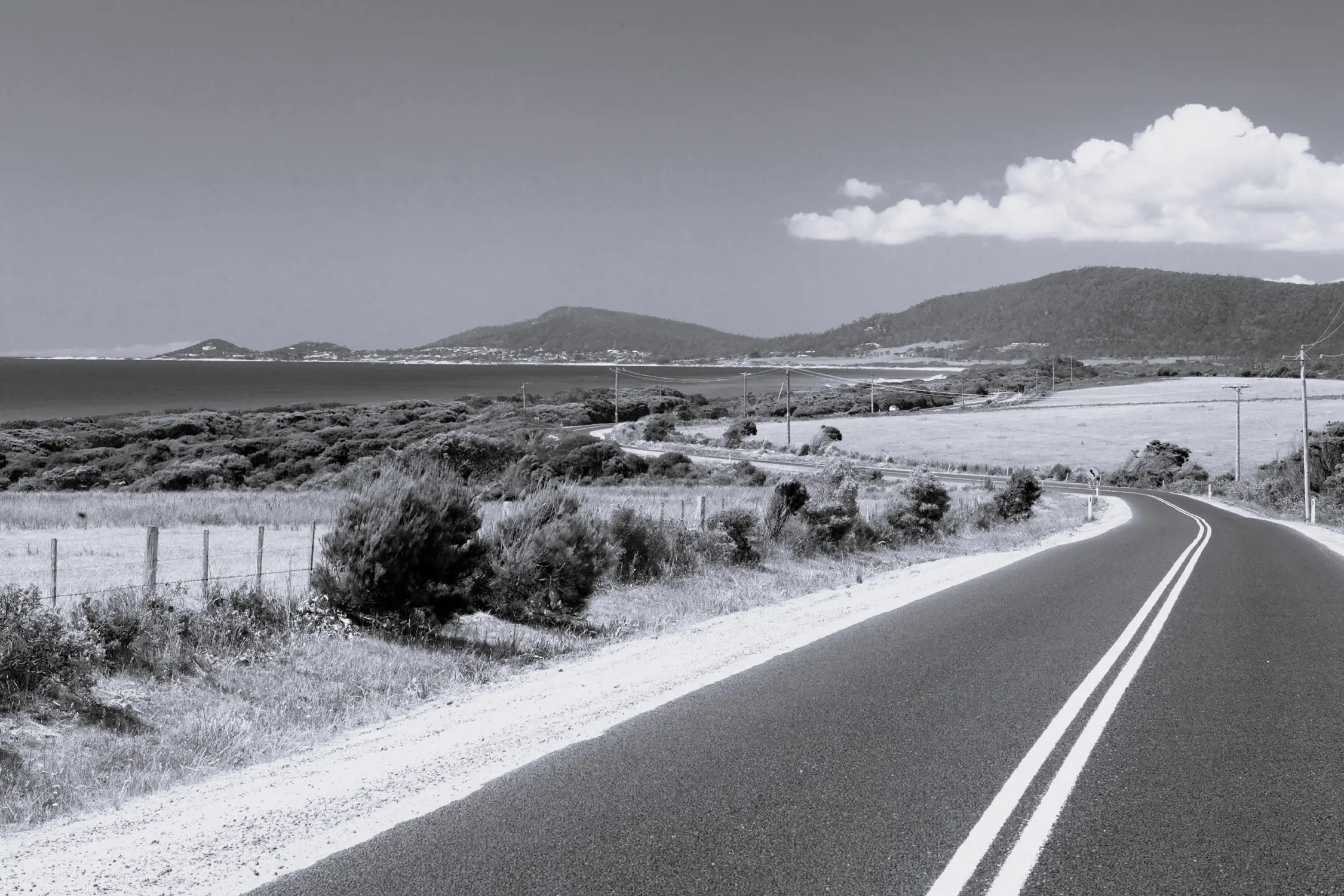 Black and white photo of a winding road running through a rural landscape with fields, bushes, power lines, and hills in the distance. The road has double white lines in the center.