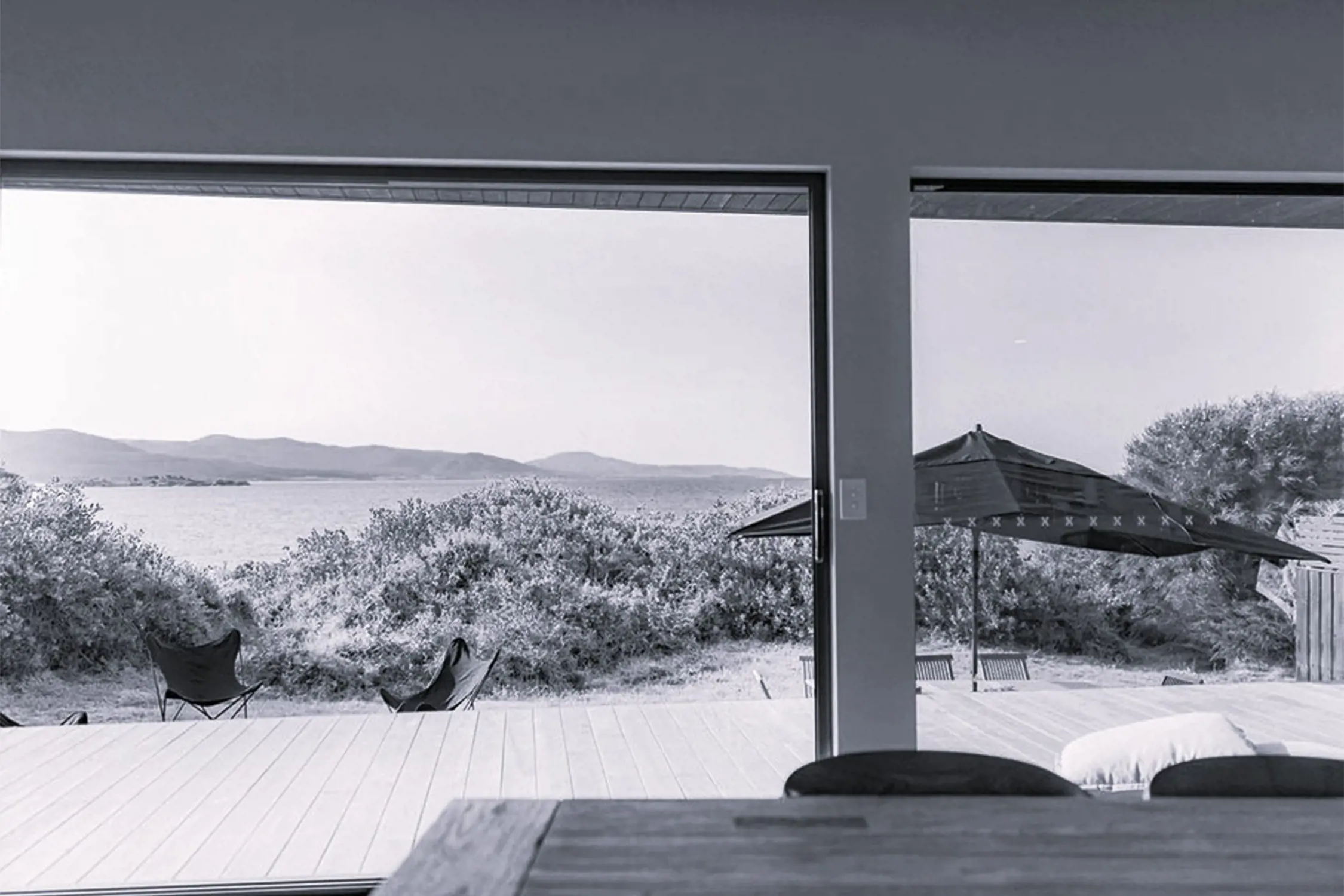Black and white photo taken from inside a room looking out through large glass sliding doors onto a deck with outdoor chairs and an umbrella, overlooking bushes and a body of water with land in the distance. A table and cushions are partially visible inside.