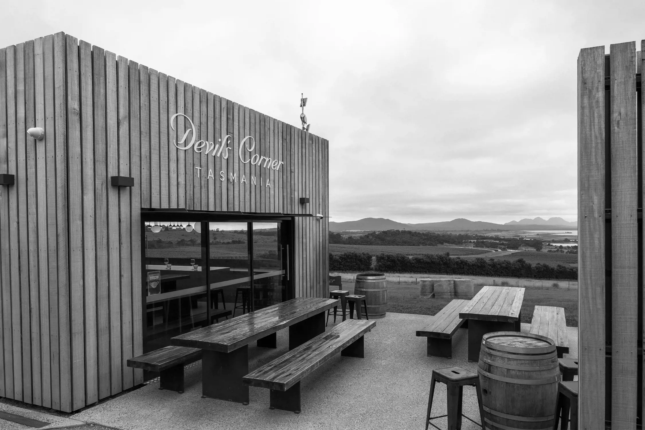 Black and white photo of an outdoor seating area at Devils Corner Tasmania vineyard, featuring wooden picnic tables, stools, barrels, and a fence overlooking vineyards and distant hills.