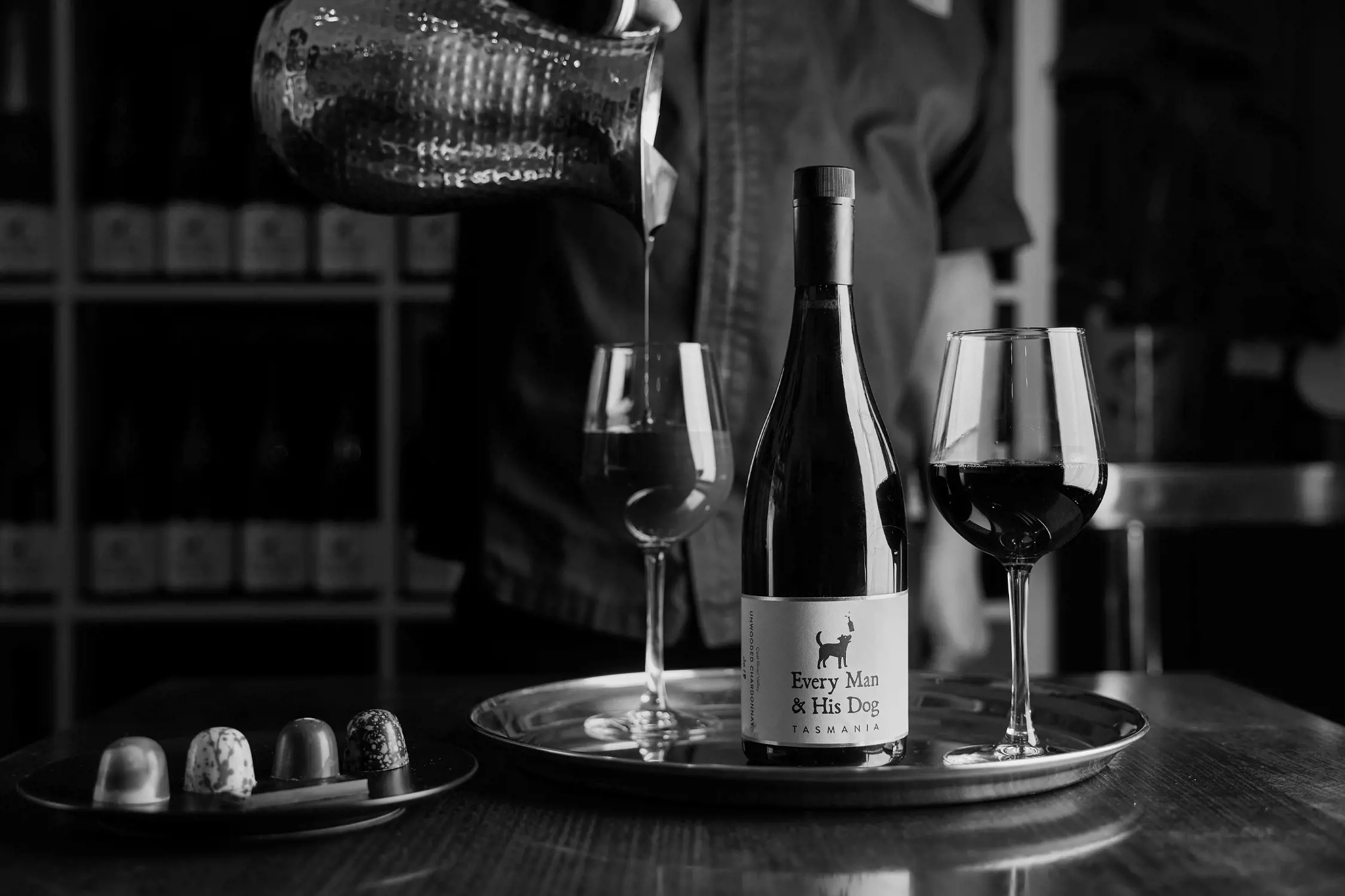 Black and white close-up photo of a bottle of Every Man & His Dog Tasmania wine on a tray alongside two glasses of red wine and a small plate of assorted chocolates, with a person pouring wine in the background.
