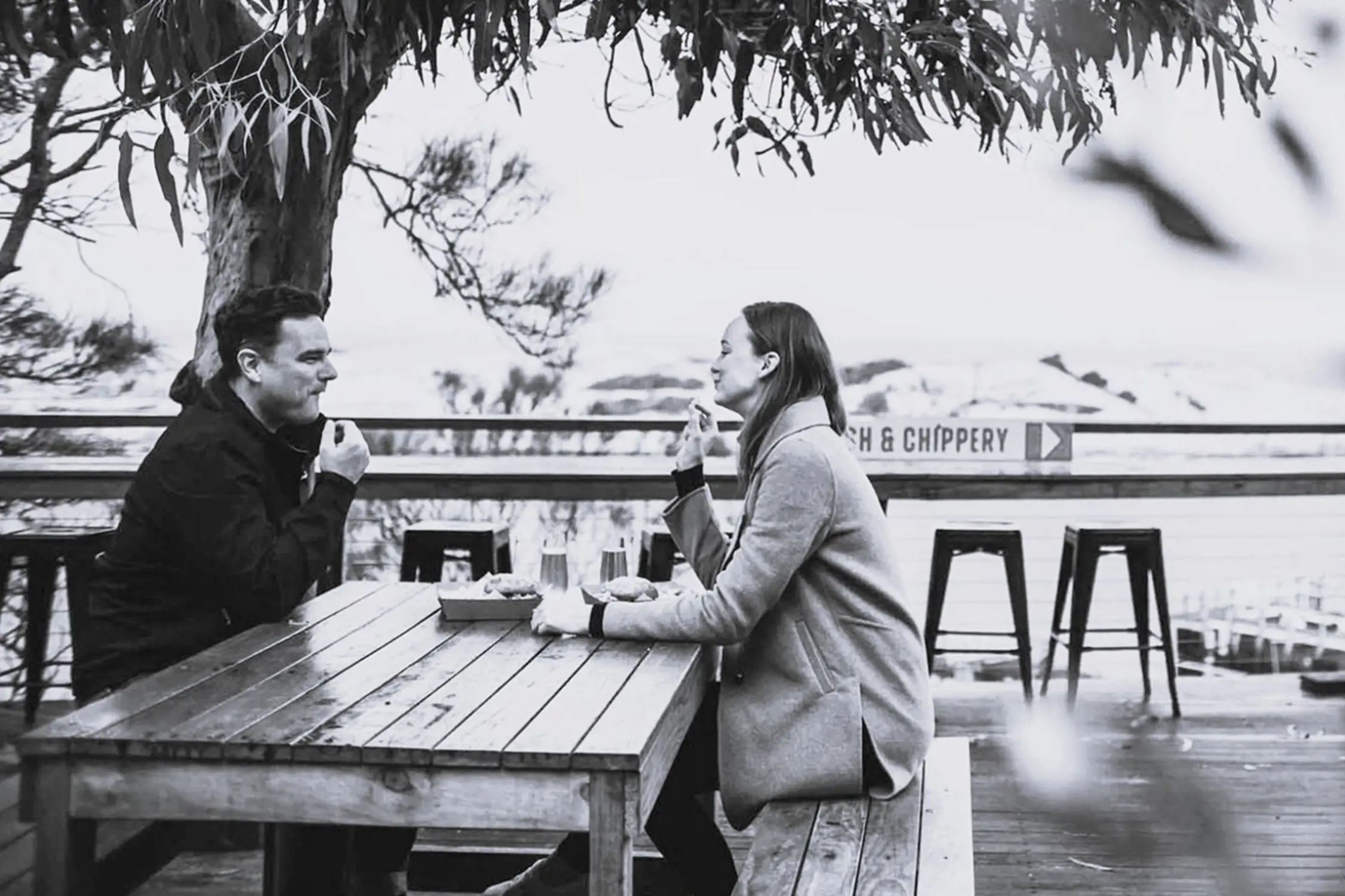 Black and white photo of a man and woman sitting at a wooden picnic table outdoors under a tree, facing each other and eating food with a calm background view of water and trees.