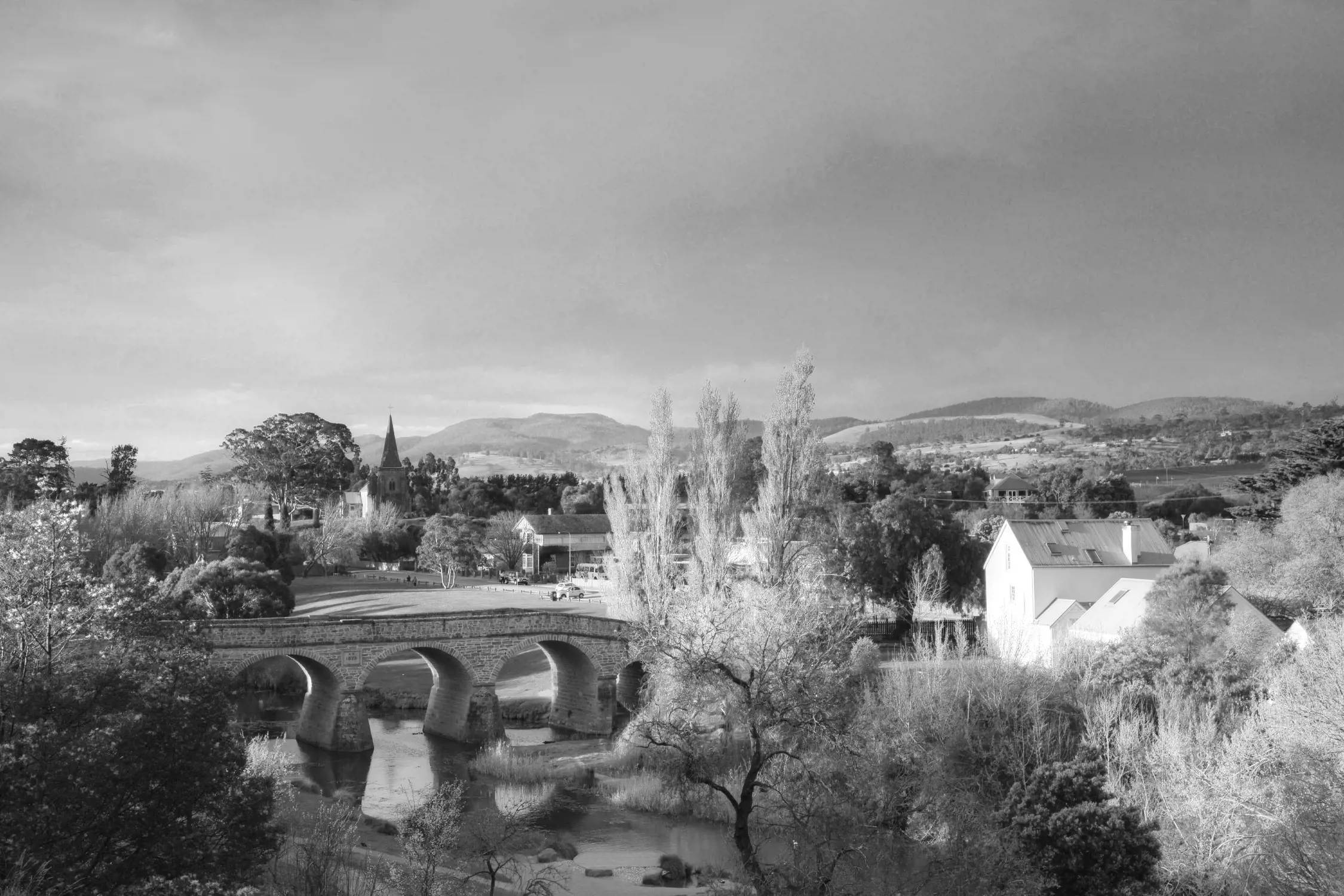 Black and white aerial photo of a historic stone bridge spanning a river in a small town, with trees, buildings, and hills visible in the background.