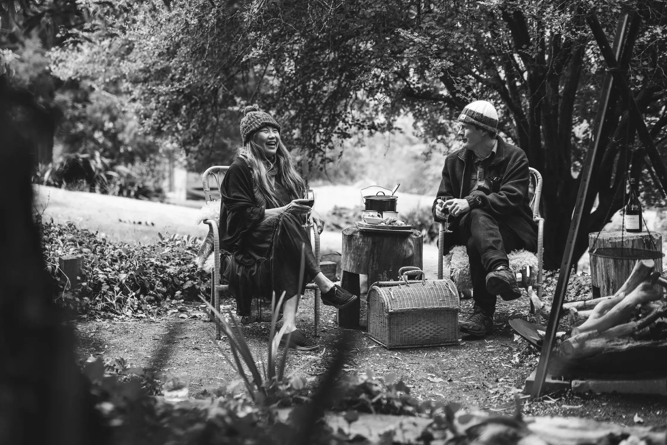 Black and white photo of two people sitting outdoors on chairs in a garden-like setting, smiling and conversing near a small table with food and a pot, surrounded by trees and nature.