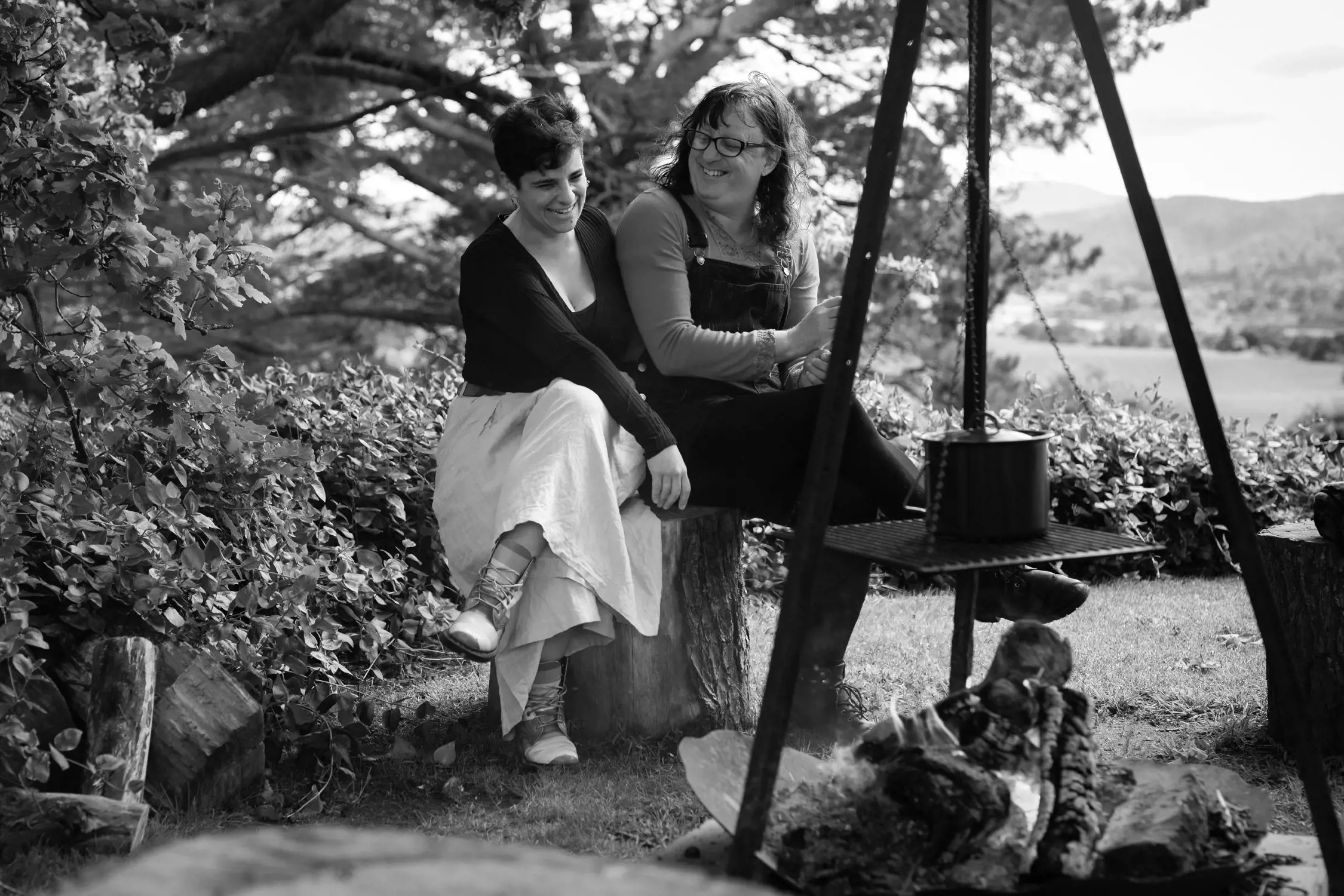 Black and white photo of two women sitting outside near a campfire, laughing and enjoying each other's company, surrounded by grass, bushes, and trees.