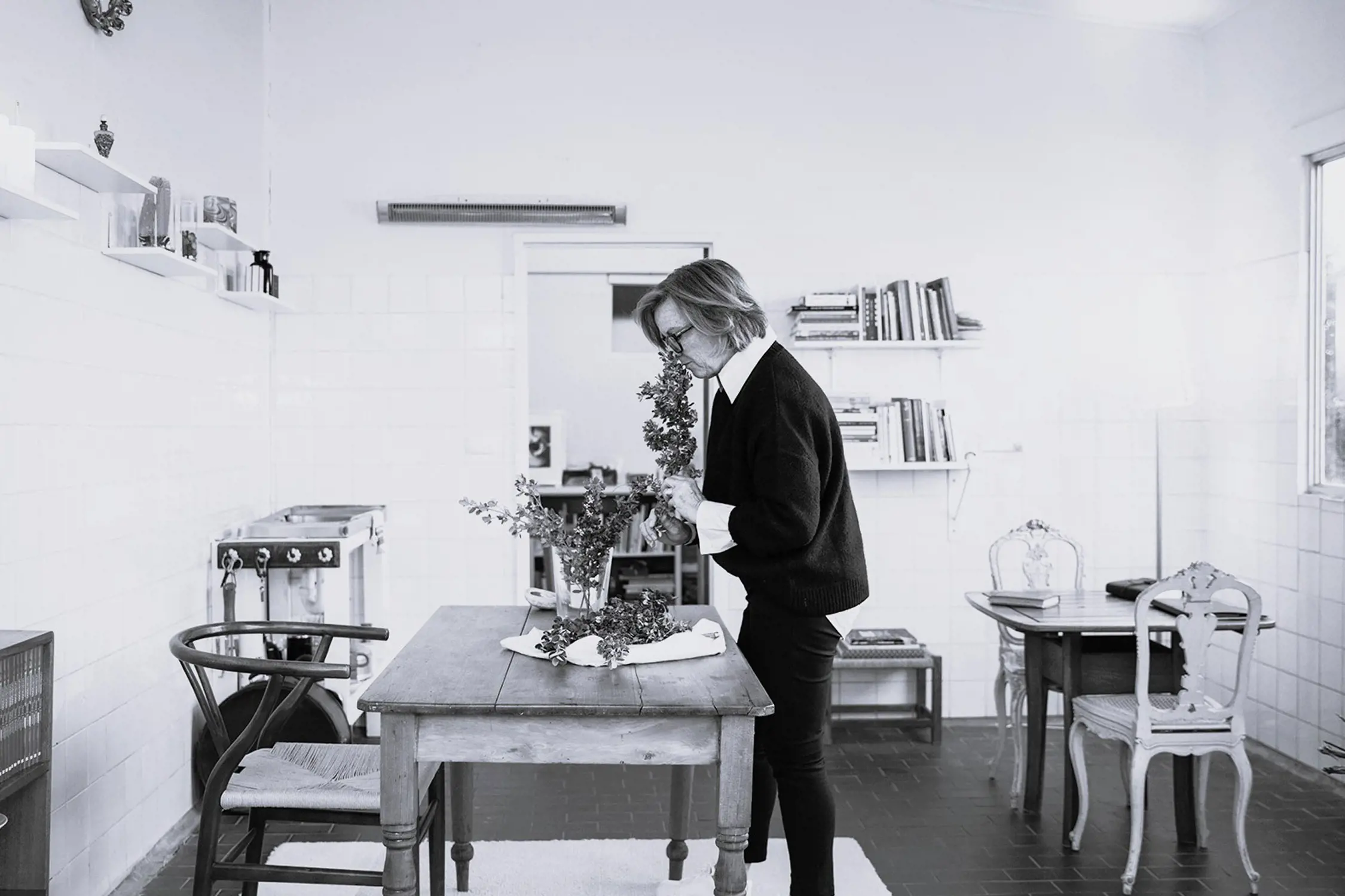 Black and white photo of a woman standing in a bright room, arranging or smelling flowers on a wooden table surrounded by chairs, shelves with books, and a window letting in natural light.