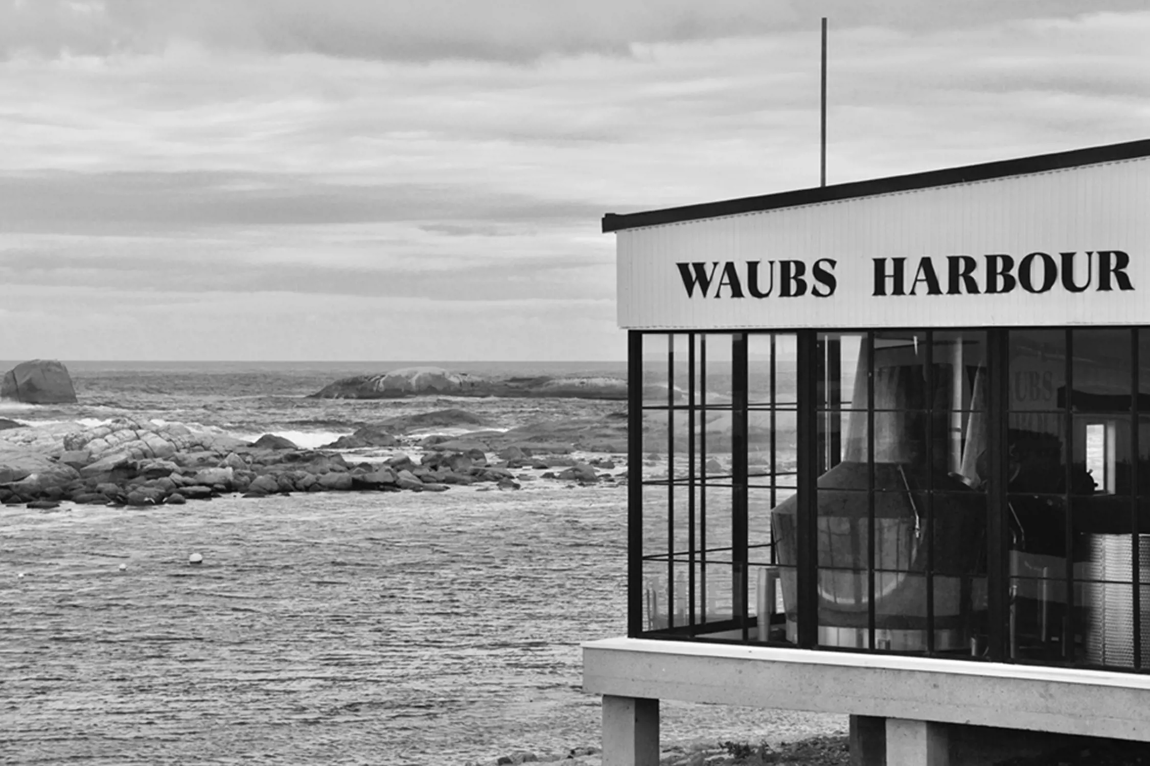 Black and white photo of a building labeled "Waubs Harbour" overlooking a rocky coastline with waves crashing and the ocean stretching to the horizon under a cloudy sky.