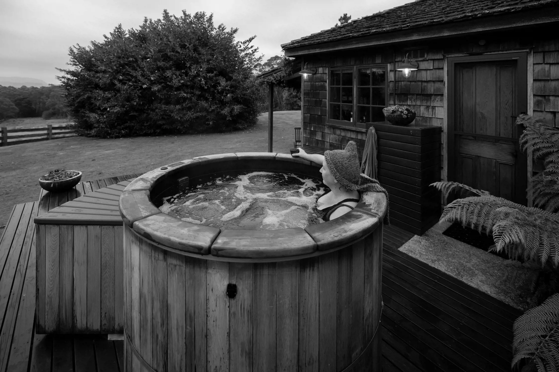 On a wooden deck beside a rustic cabin, a person wearing a knitted hat relaxes in a round, wooden hot tub filled with bubbling water. The individual leans back on the hot tub’s edge, holding a drink in one hand, surrounded by greenery and outdoor lighting, creating a peaceful retreat atmosphere.