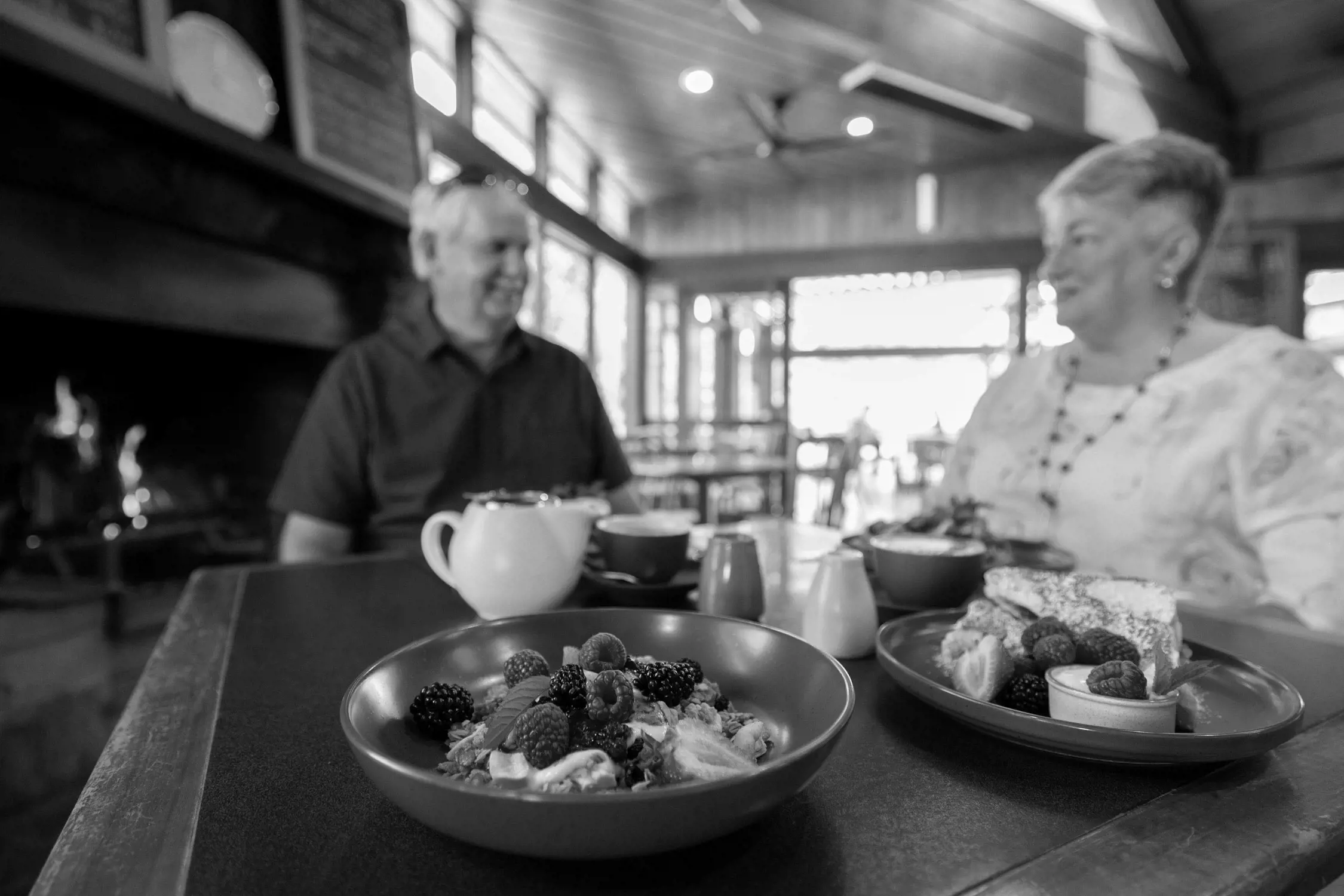 Inside a cozy restaurant with wooden interiors, an elderly man and woman sit facing each other, smiling and engaging in conversation. The foreground features a wooden table with colorful dishes including a bowl of granola topped with fresh blackberries and raspberries, a plate with pastry and fruit, a white teapot, and cups of coffee or tea.