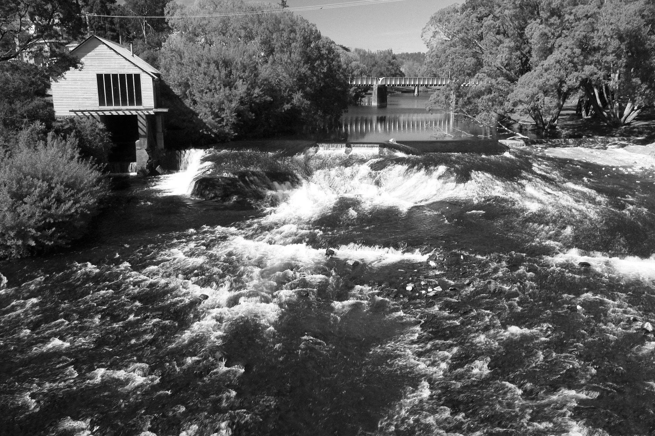 A wide river cascades over a series of rock ledges, creating a low waterfall with flowing white water and ripples. Above the waterfall, a steel truss bridge stretches across the river, framed by lush trees and dense forest hillsides reflected on the calm water’s surface in the foreground.