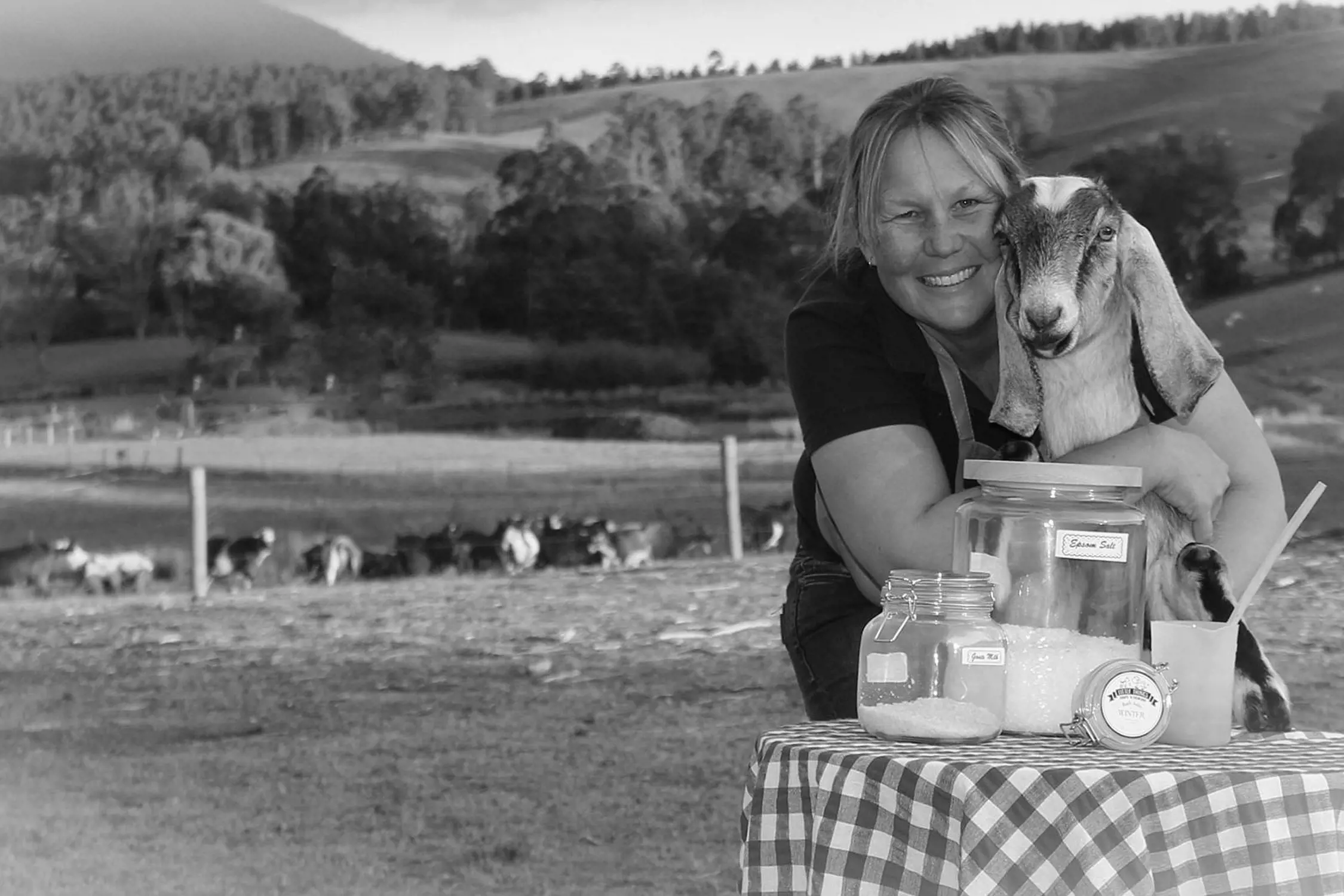 A cheerful woman with light hair pulled back smiles warmly while holding a young goat close to her chest in a rural farm setting. She stands behind a checkered tablecloth-covered table displaying several large glass jars filled with labeled salts and bath products, with green pastures and distant trees in the background under bright daylight.