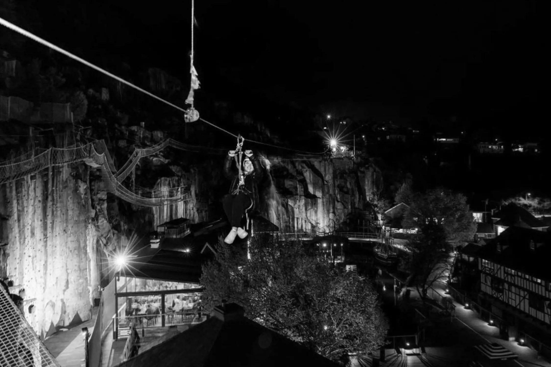 Person riding a zip line at night over a small town, with illuminated buildings and rocky cliffs in the background.