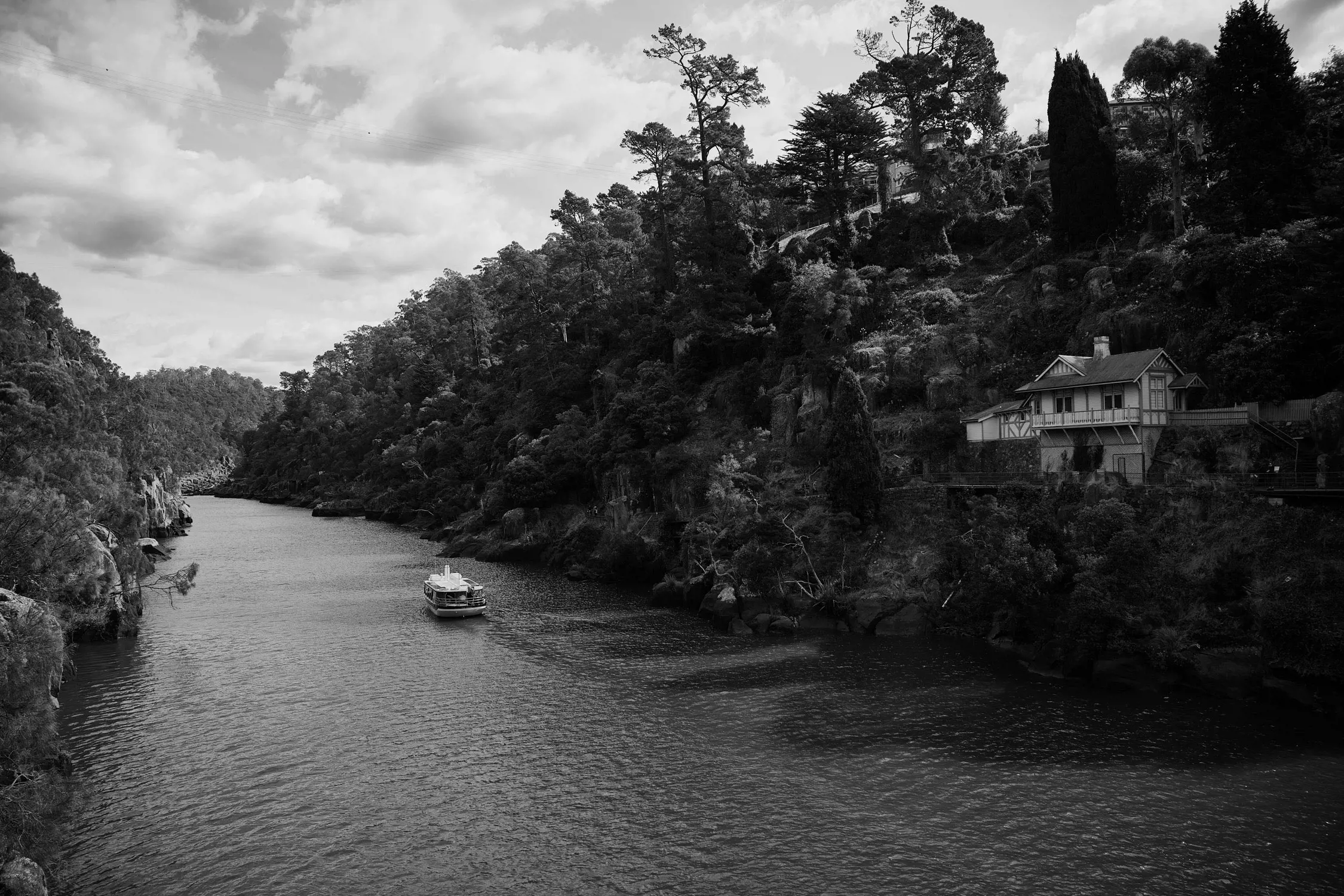 Serene river flowing between wooded banks, with a boat on the water and a house perched on the right hillside under a cloudy sky.