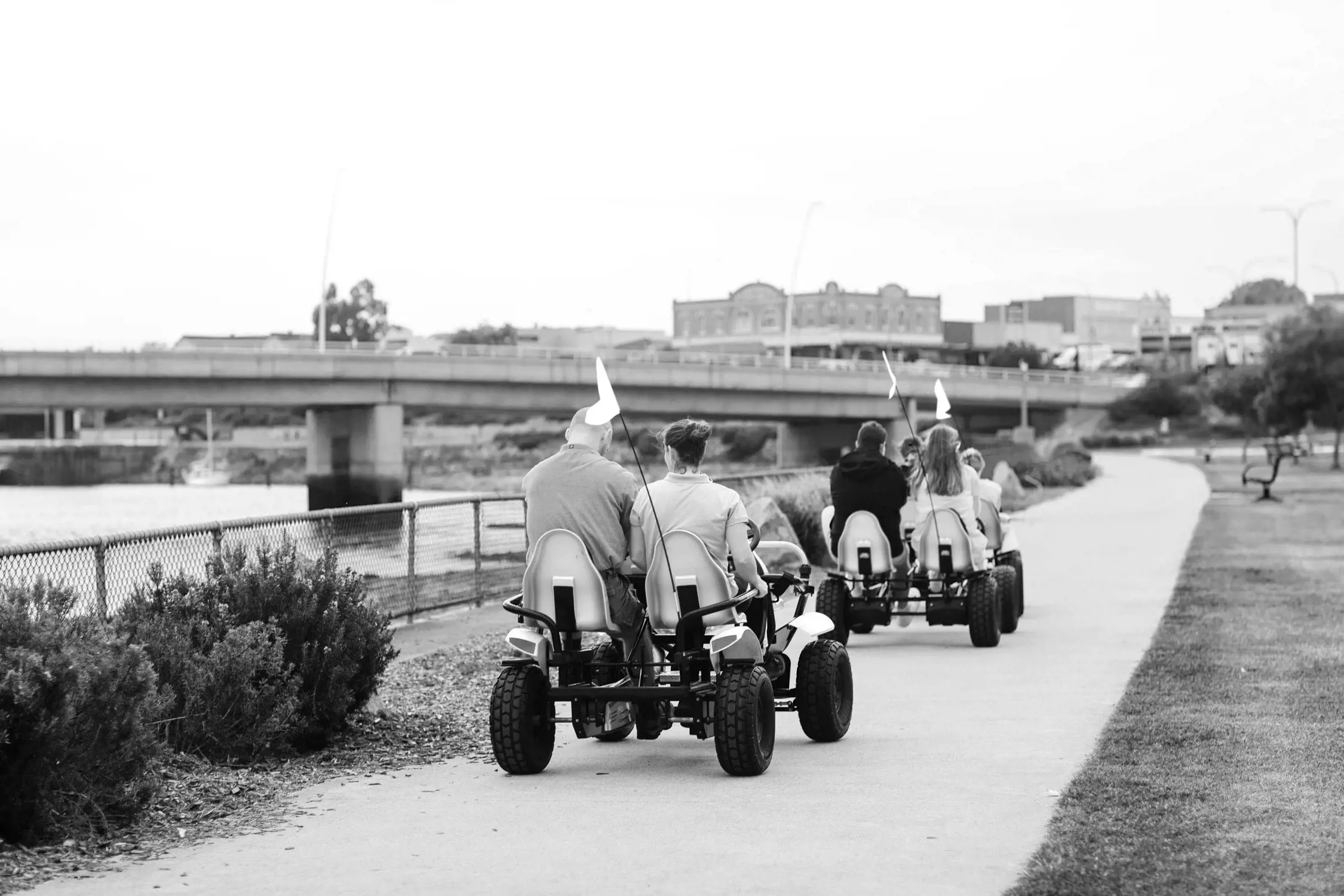 Black and white photo of two couples riding pedal-powered four-wheeled bikes along a pathway beside water, with a bridge and buildings in the background.