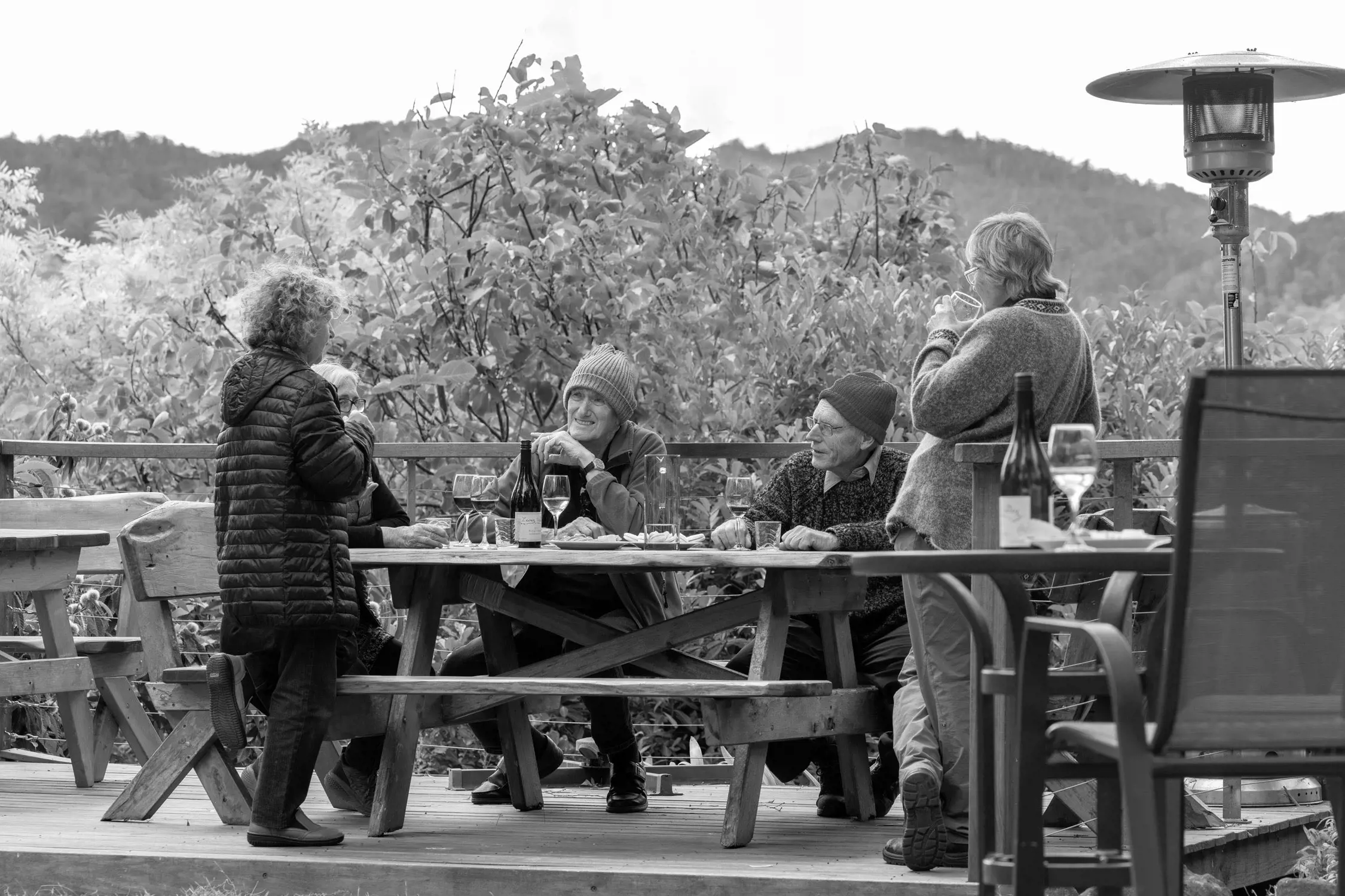 Black and white photo of a group of older adults enjoying drinks and conversation at a picnic table on a wooden deck with a backdrop of trees and hills.