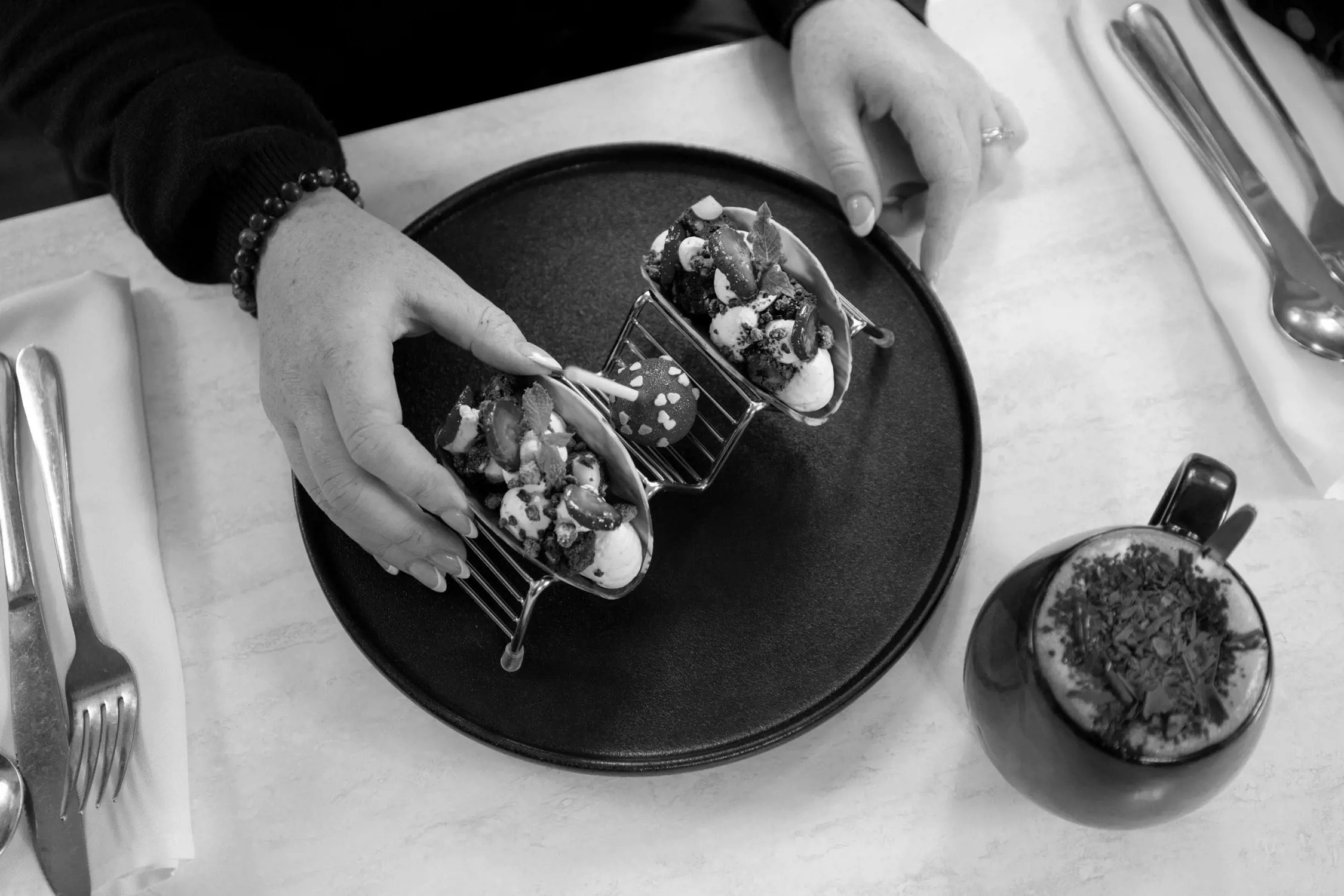 A black and white photo showing hands holding two dessert tacos filled with cream and garnished with strawberries and mint leaves on a plate, alongside a cup of hot beverage topped with whipped cream and chocolate shavings.