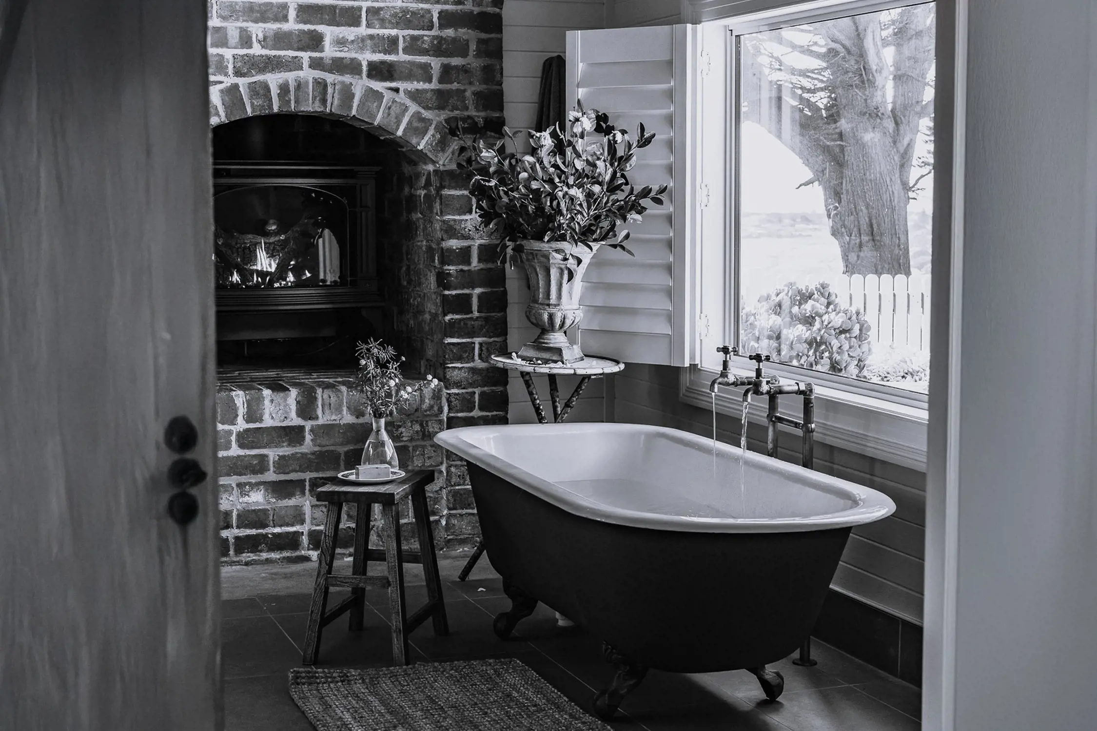 A black and white image of a vintage-style bathroom featuring a clawfoot bathtub with running water, a brick fireplace, a small wooden stool with a vase and soap, and a window showing outdoor greenery.