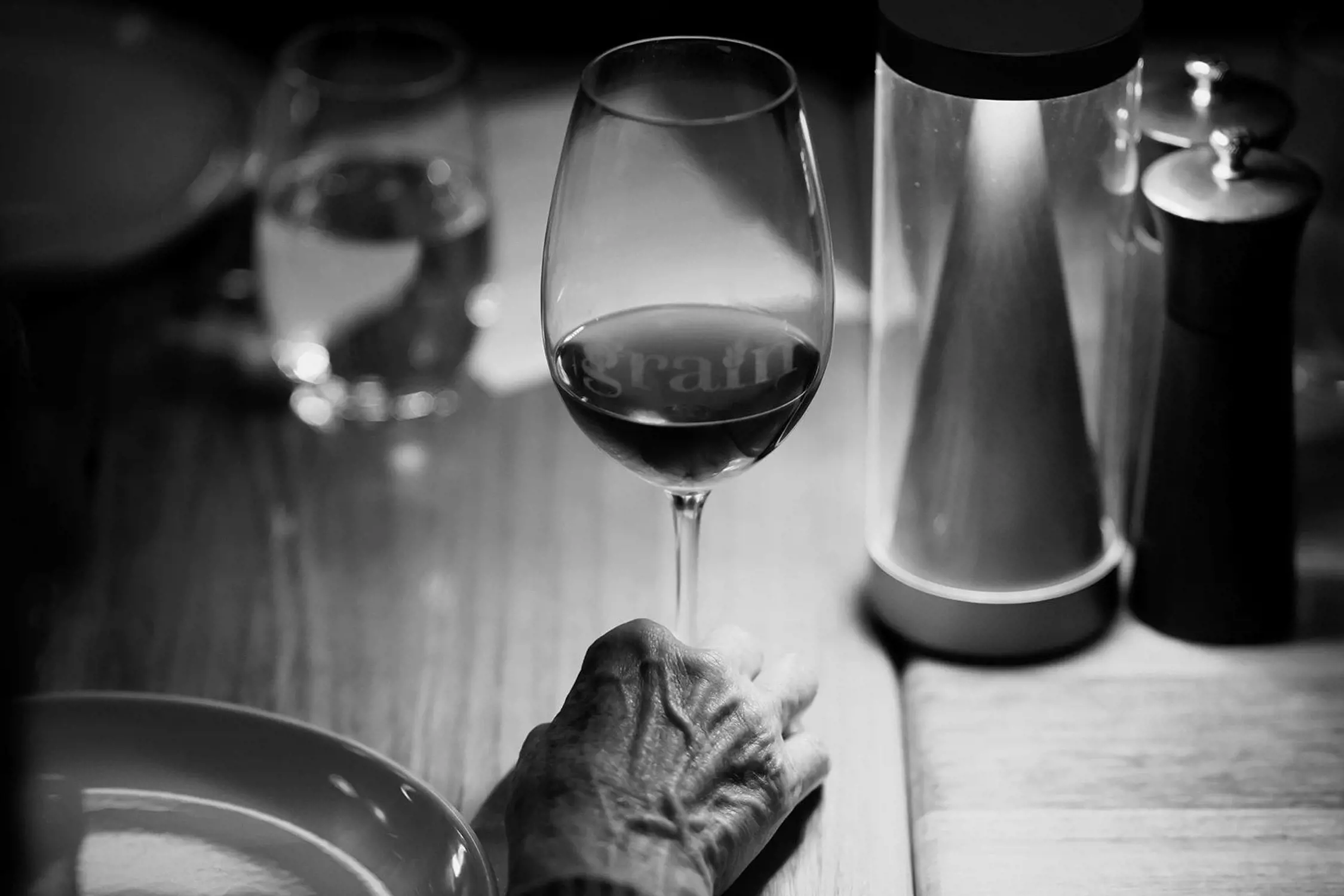 An elderly hand resting on a wooden bar counter next to a glass of red wine, with various bottles and bar items blurred in the background.