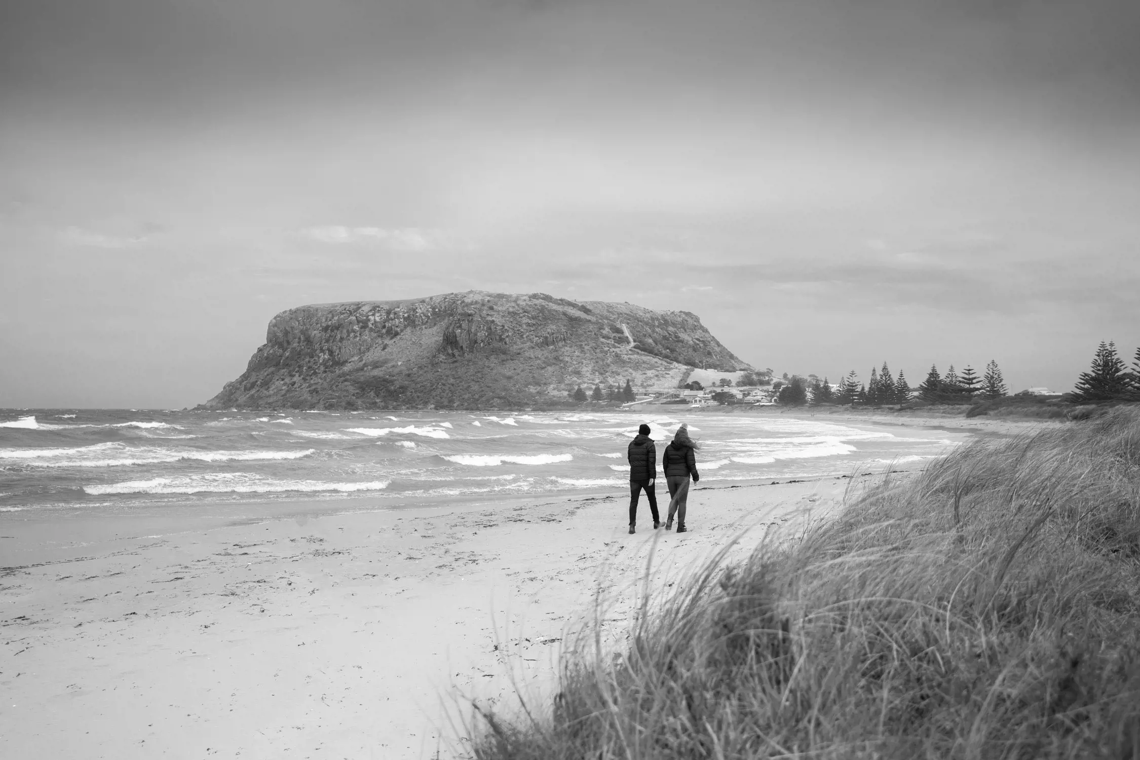 A black-and-white wide shot of two people walking along a sandy beach with waves crashing to the left and a large rocky mountain in the background; tall grass and trees line the sides of the beach.