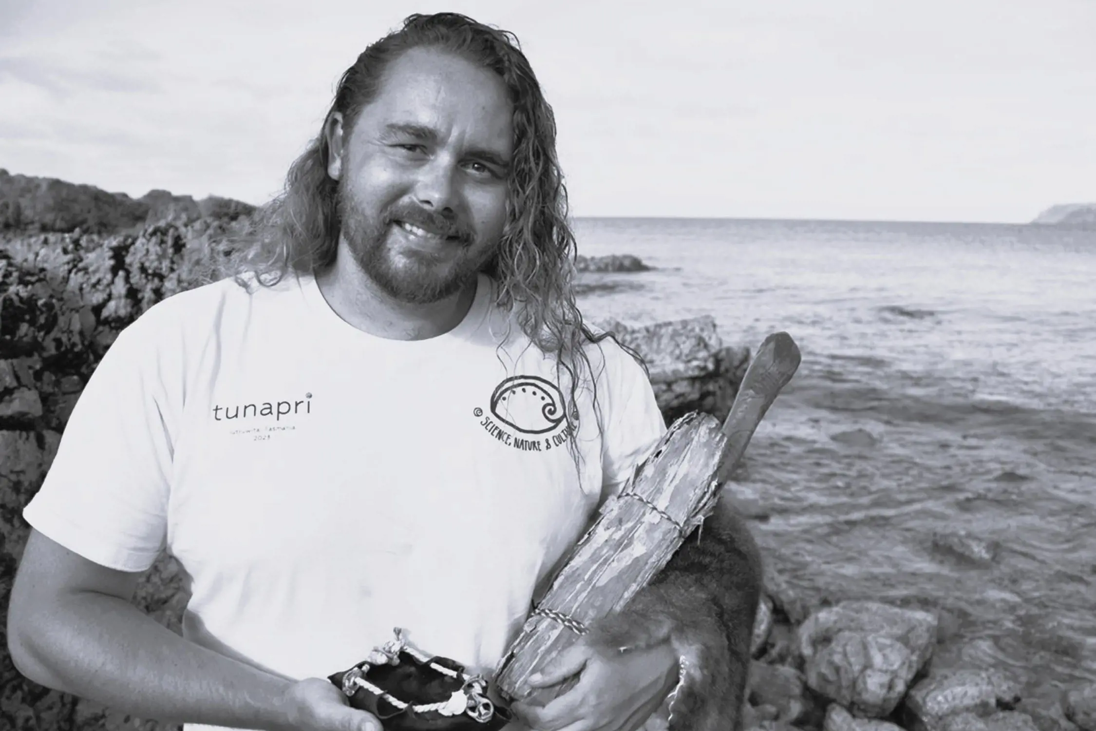 A black-and-white image of a man with long hair and a beard standing by a rocky shoreline, smiling and holding traditional cultural objects, with the sea and sky visible behind him.