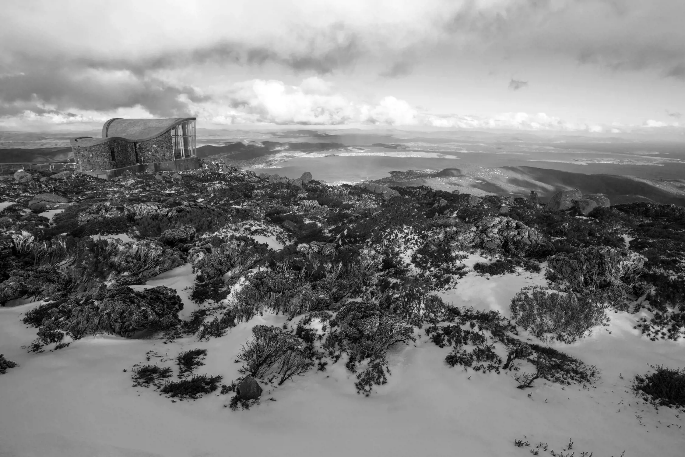 Black and white panoramic view from a mountaintop showing a modern building with a curved roof, rocky terrain with patches of snow, vegetation, and distant valleys under a cloudy sky.