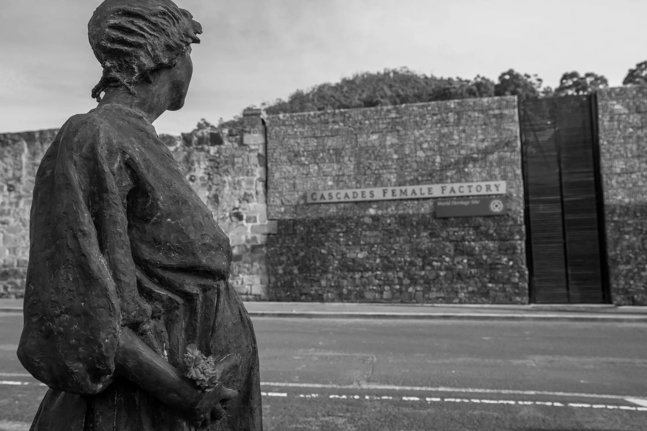 Black and white photo of a bronze statue of a woman holding flowers, looking towards the Cascades Female Factory World Heritage site building made of stone.