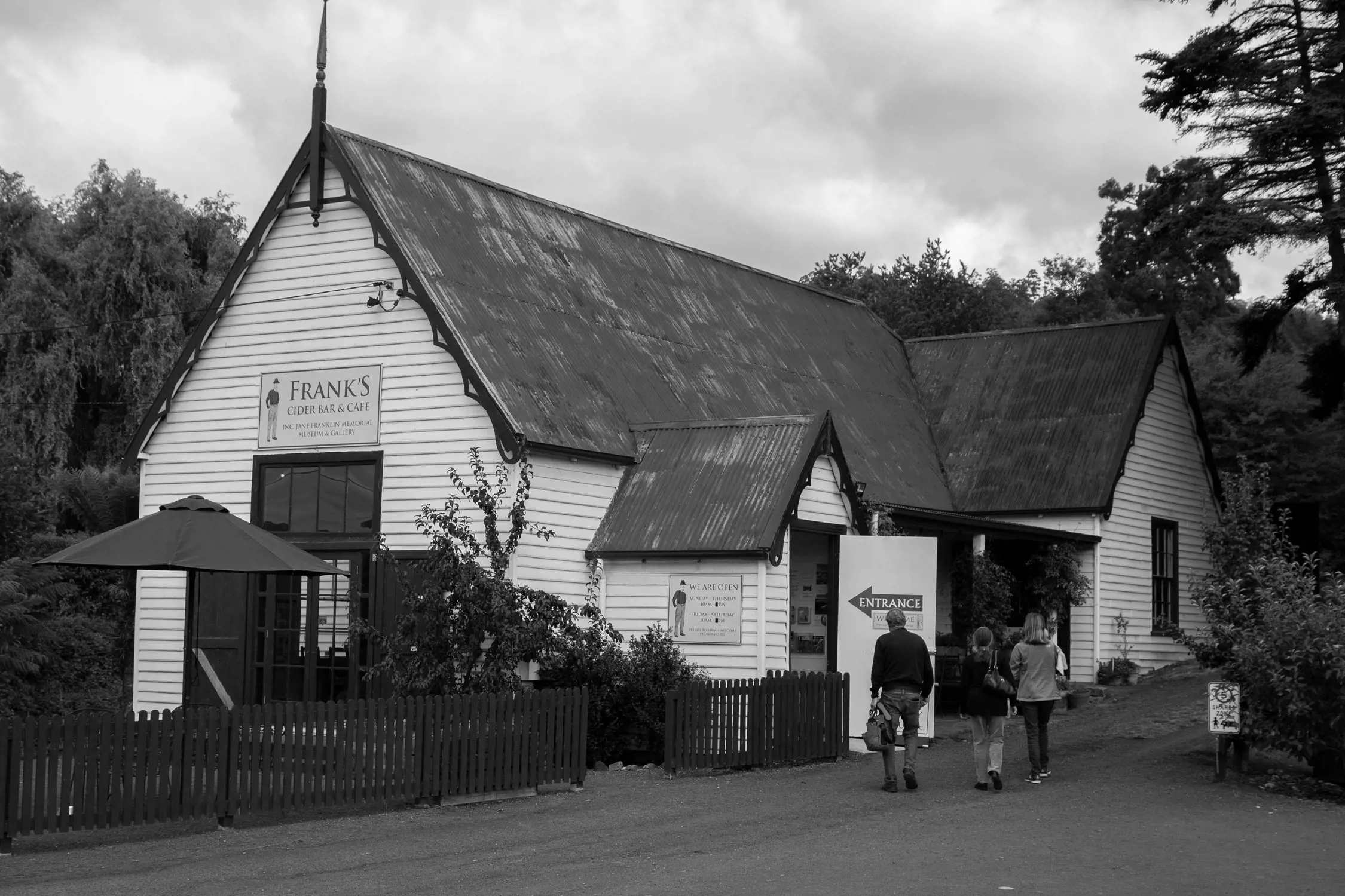 Black and white photo of a historic white wooden building labeled "Frank's Cider Bar & Cafe" with people walking toward the open entrance.